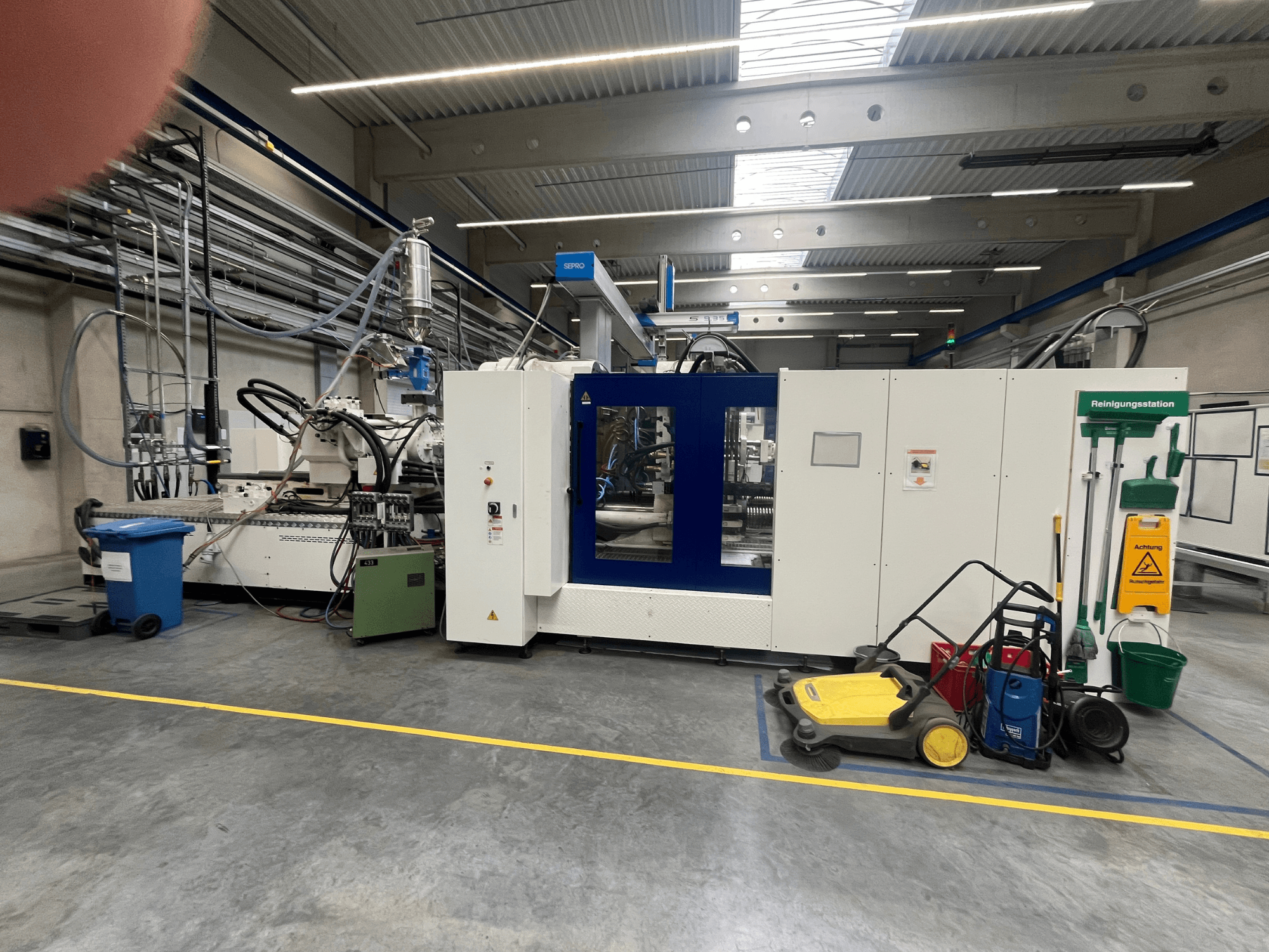 Máquina industrial numa fábrica, vista de frente, com painéis azuis e brancos, mangueiras e equipamento de limpeza nas proximidades.