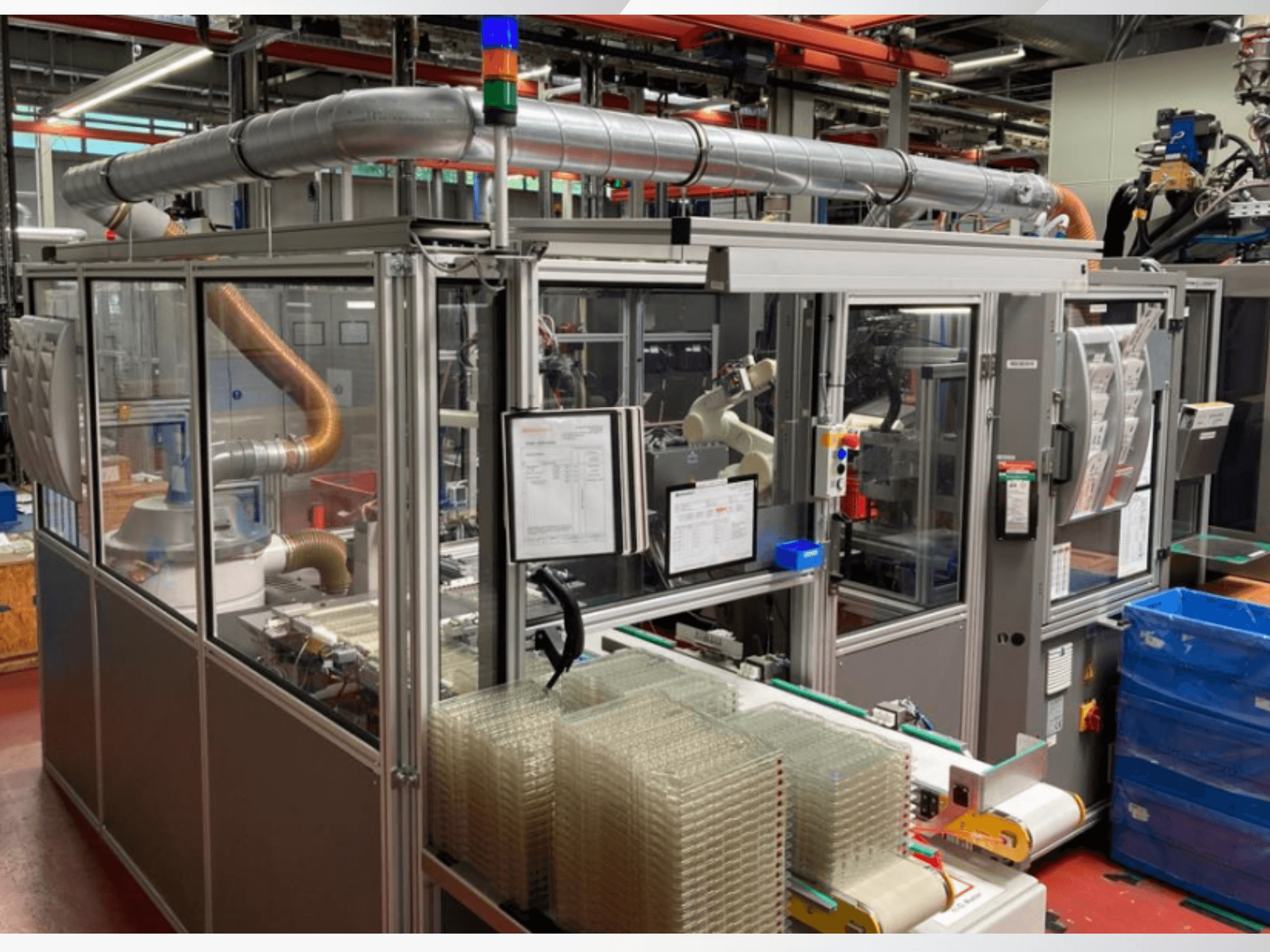 Vista frontal de uma máquina automatizada com tubos de metal, painel de controlo e recipientes de plástico transparente empilhados no chão de uma fábrica.