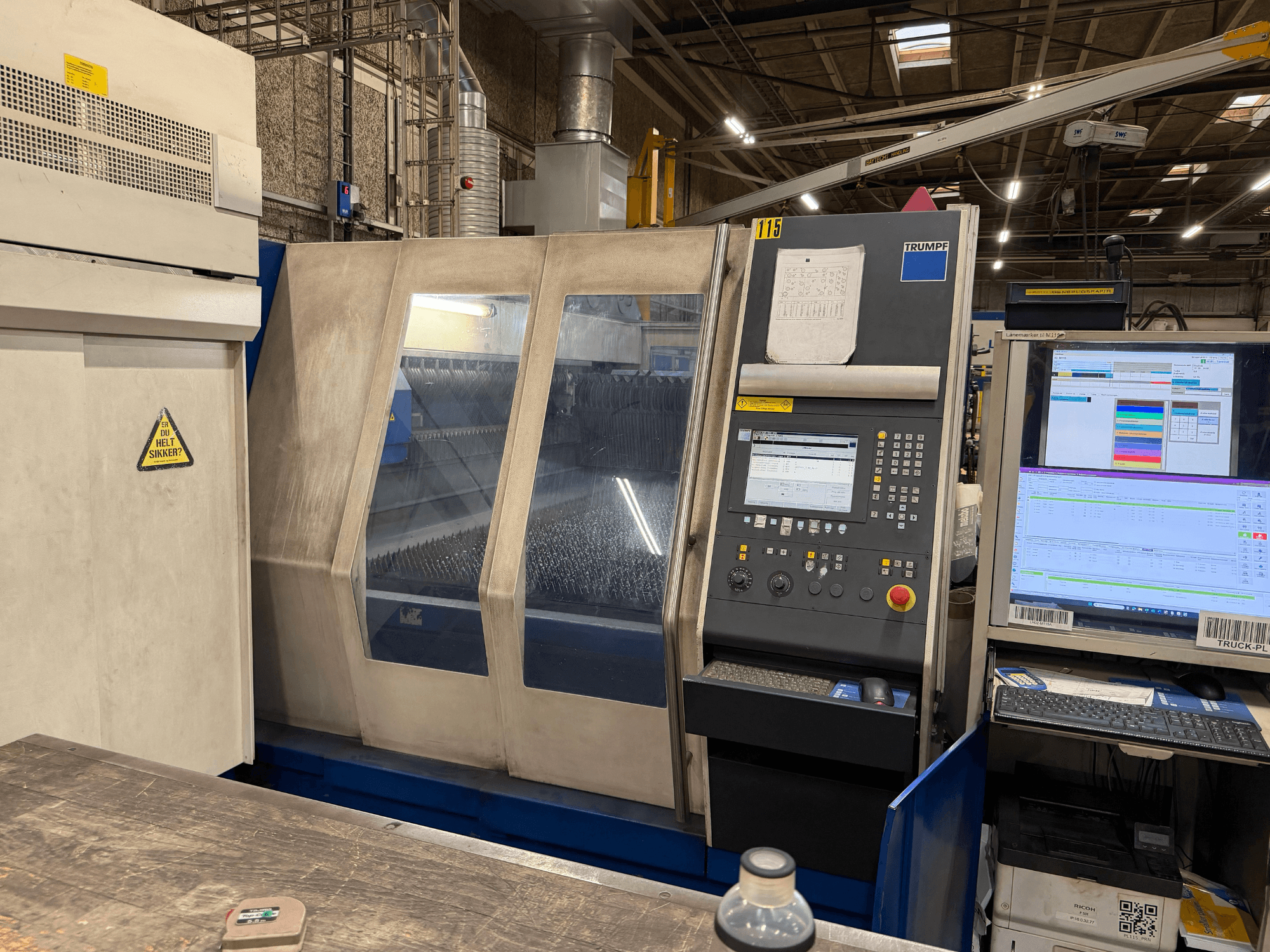 Vista frontal de uma máquina laser industrial Trumpf com painel de controlo e ecrãs digitais, colocada num ambiente de fábrica.