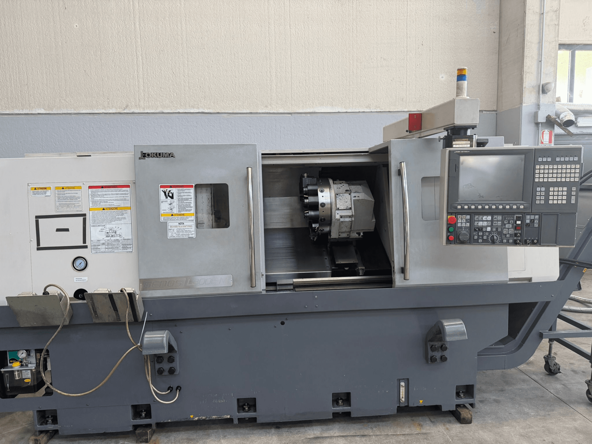 Torno CNC Okuma, vista frontal, mostrando o painel de controlo, o suporte da ferramenta de corte e os avisos de segurança na lateral.