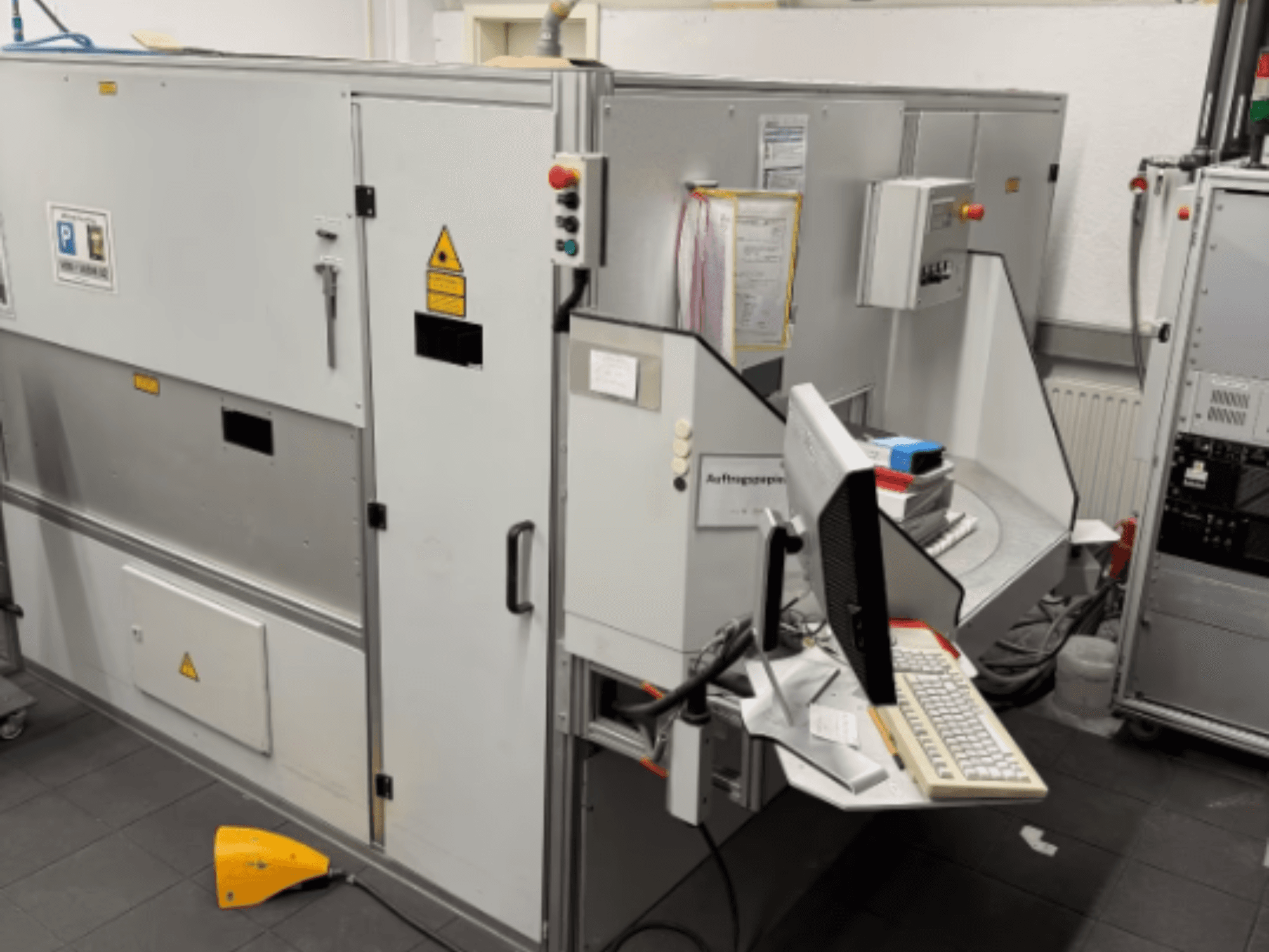 Máquina industrial com um terminal de computador, vista lateral; inclui botões de controlo e sinais de aviso de segurança.