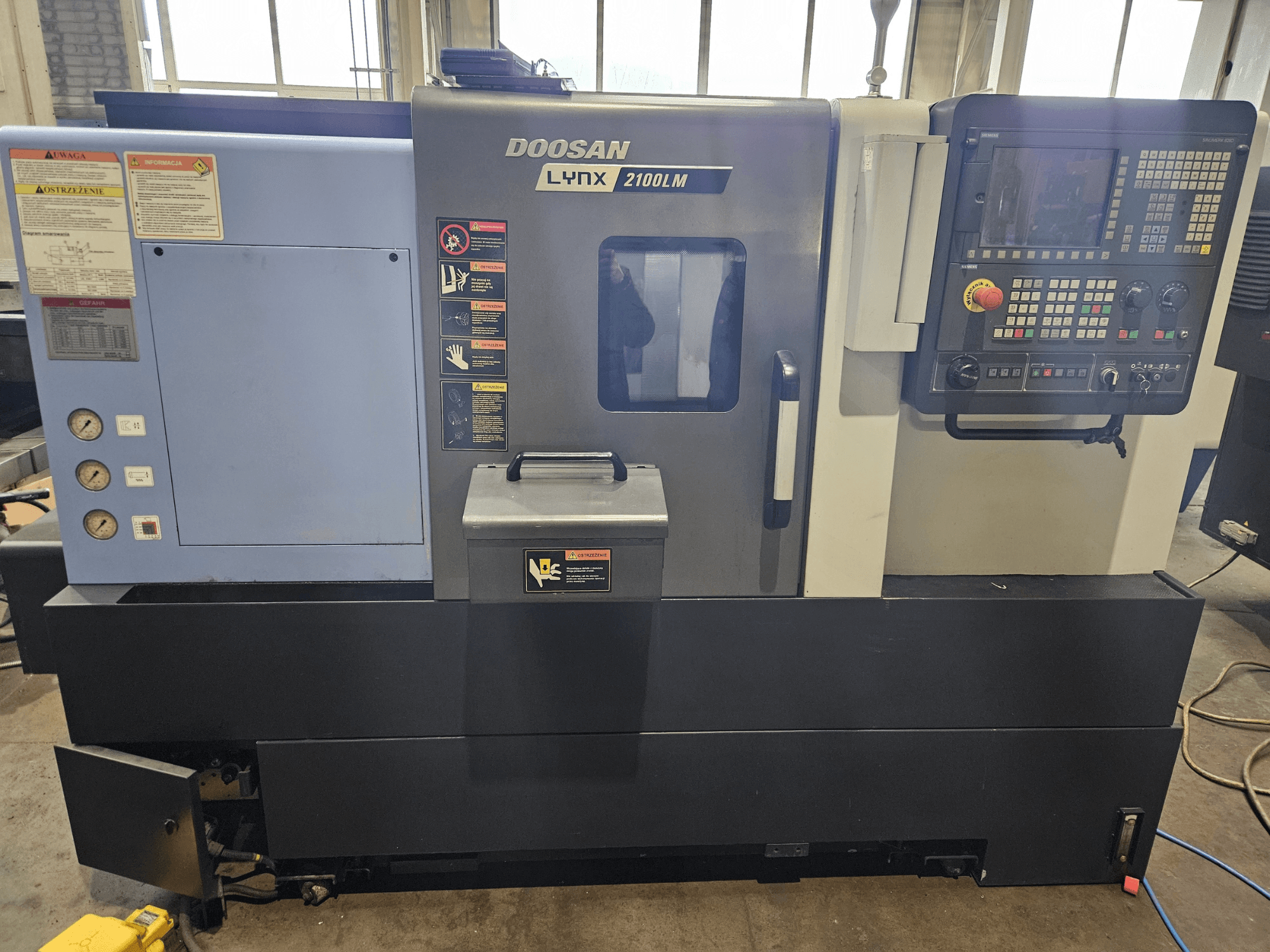 Torno CNC Doosan Lynx 2100LM, vista frontal, com painel de controlo, manómetros de pressão de ar e etiquetas de segurança visíveis.