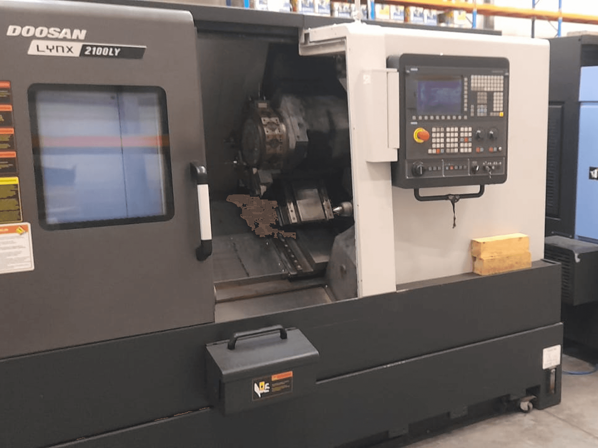 Torno CNC Doosan Lynx 2100LY, vista frontal mostrando o painel de controlo, o suporte de ferramentas e o espaço de trabalho interior; etiquetas técnicas visíveis.