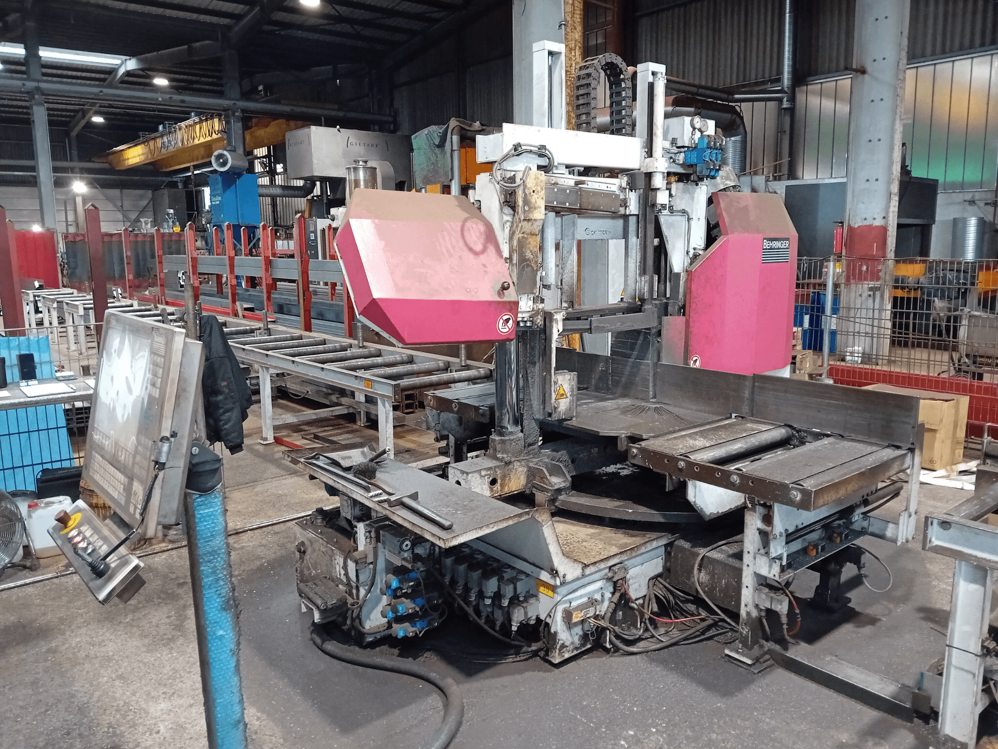 Máquina de serra de fita Behringer numa fábrica, vista frontal, com uma grande lâmina de corte e painel de controlo com botões.