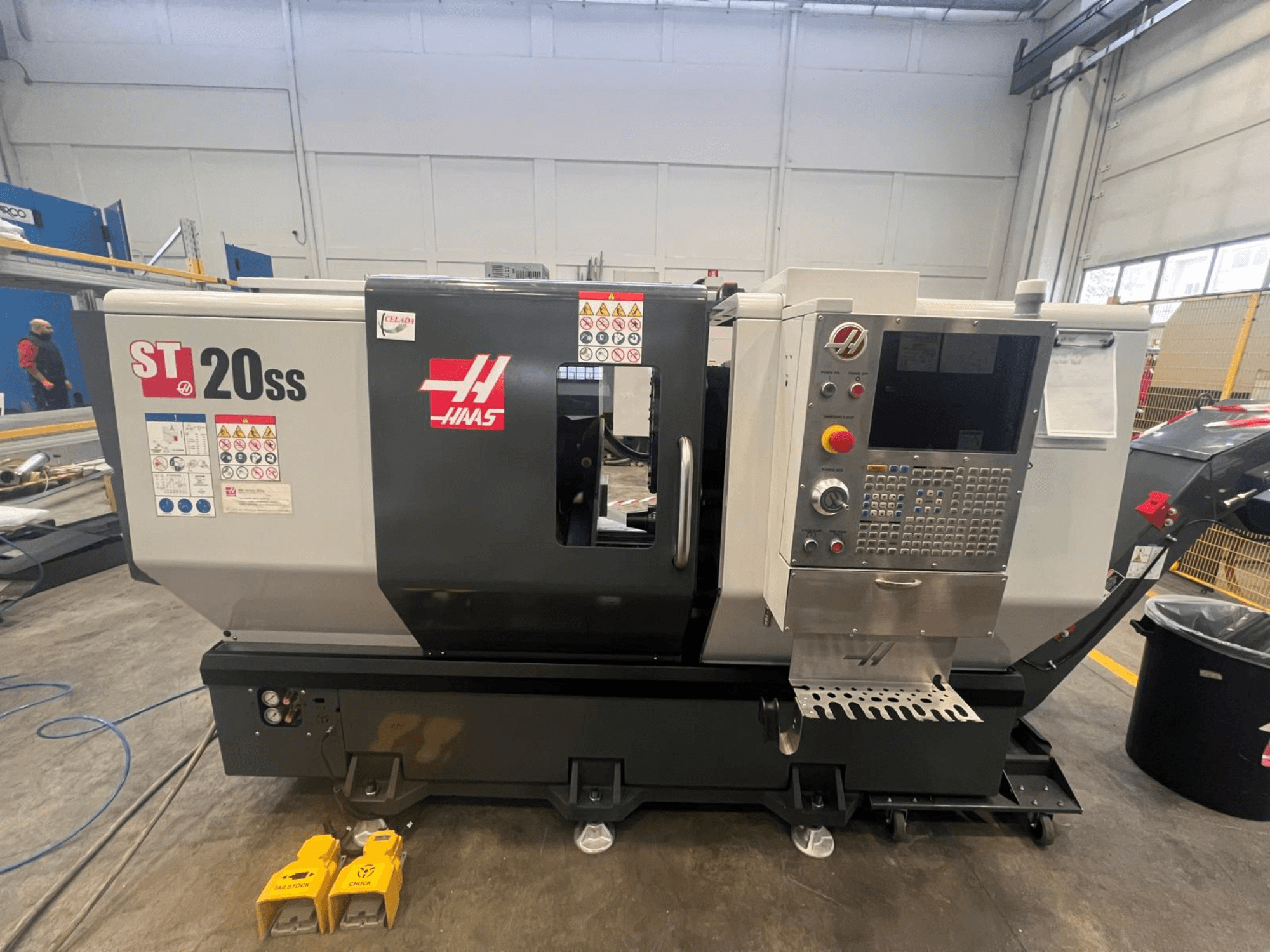 Torno CNC Haas ST 20SS em vista lateral, com painel de controlo, etiquetas de segurança e pedais no chão.