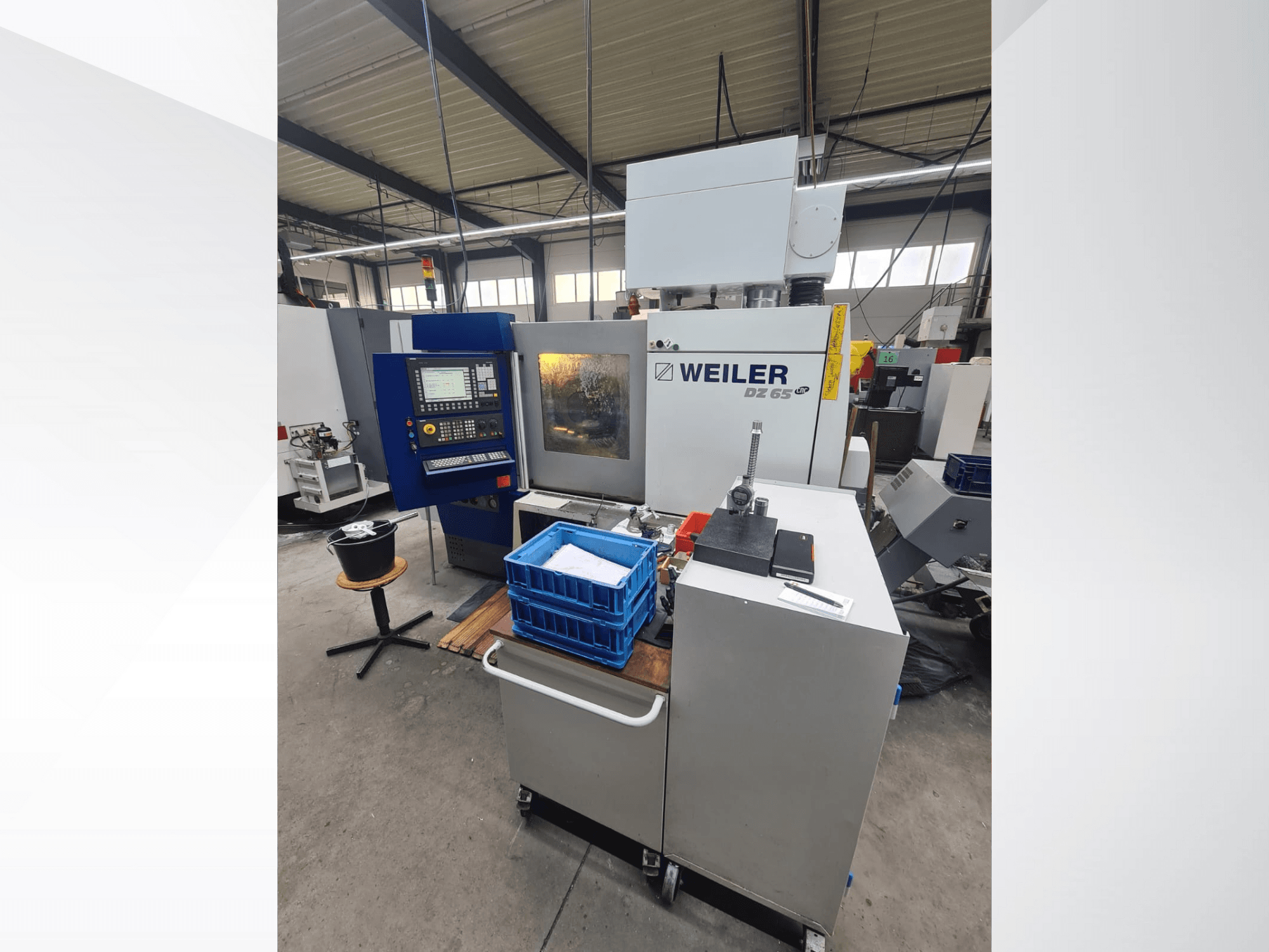 Máquina CNC Weiler DZ 65 numa oficina, vista frontal com painel de controlo, ferramentas e caixas de armazenamento azuis visíveis.