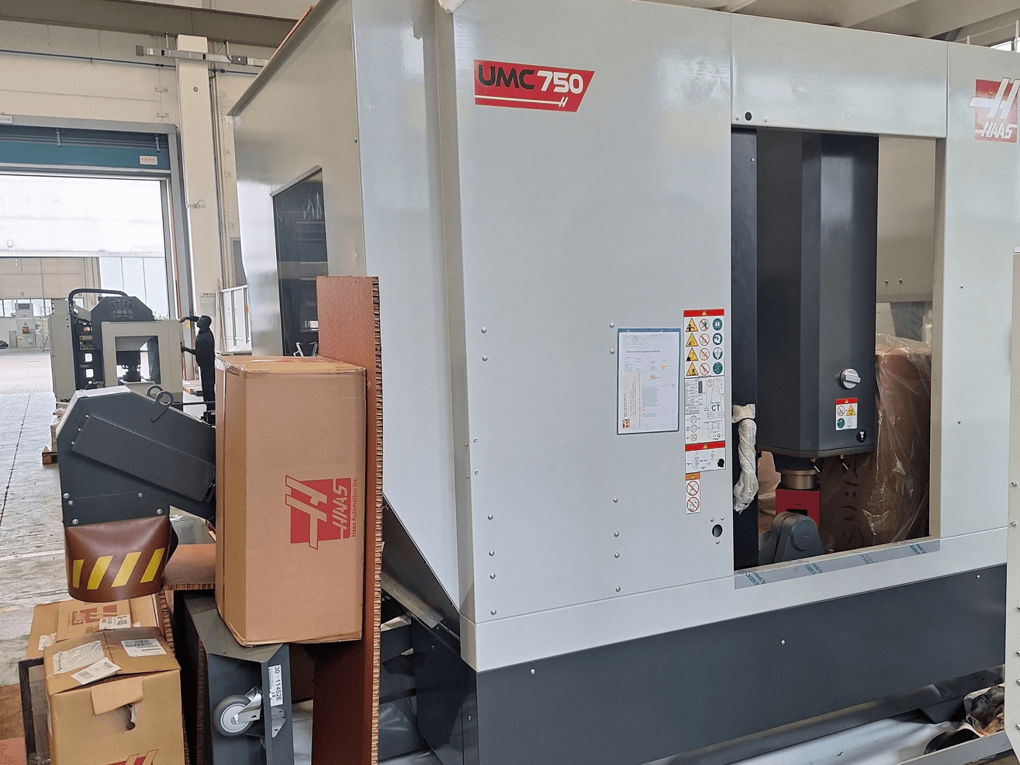 Centro de maquinagem universal HAAS UMC-750, vista lateral, mostrando o painel de controlo e o compartimento de armazenamento de ferramentas com etiquetas de aviso e embalagem.