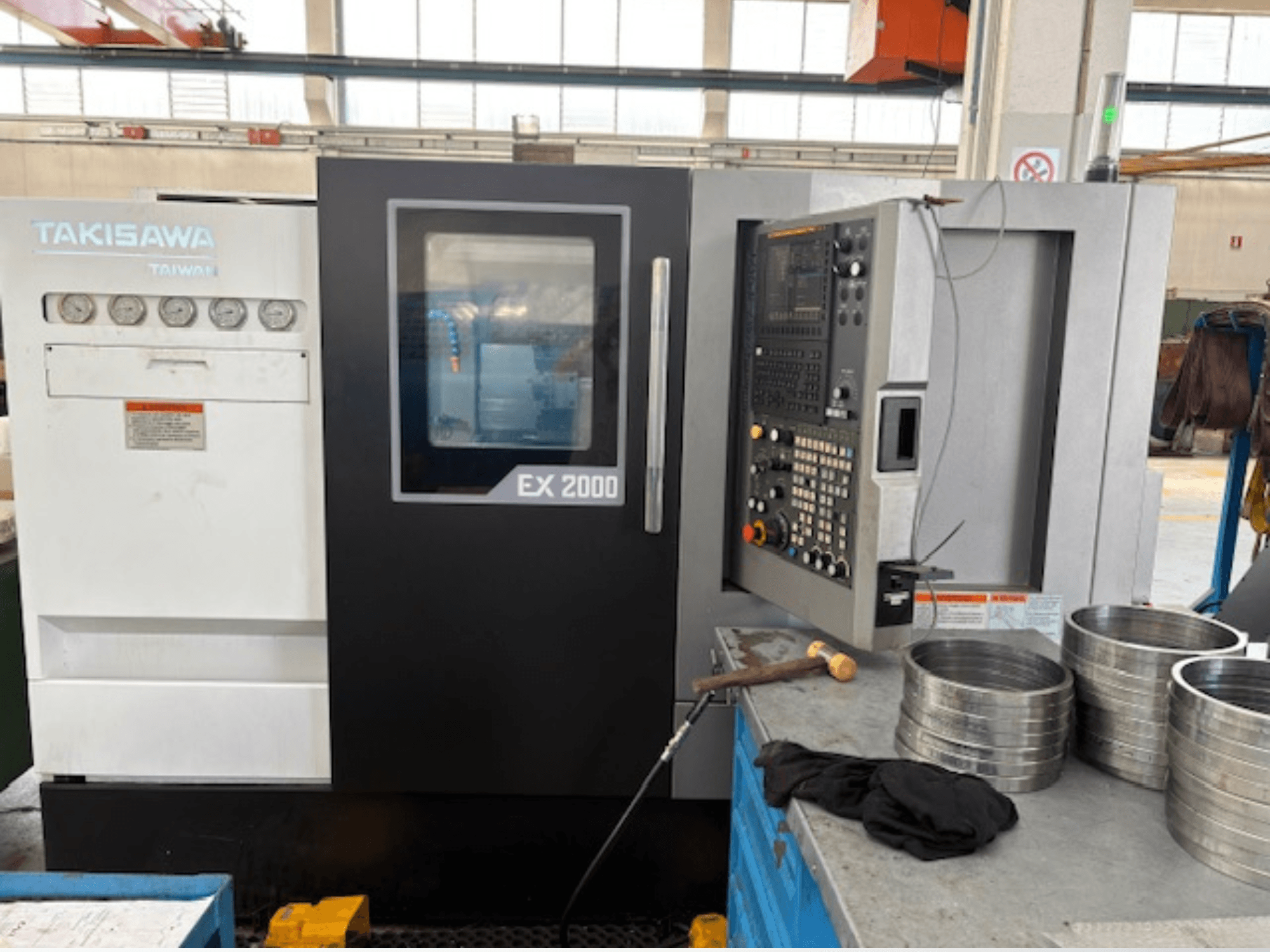 Máquina CNC Takisawa EX 2000, vista frontal, com painel de controlo, manómetros e peças metálicas numa mesa de oficina.
