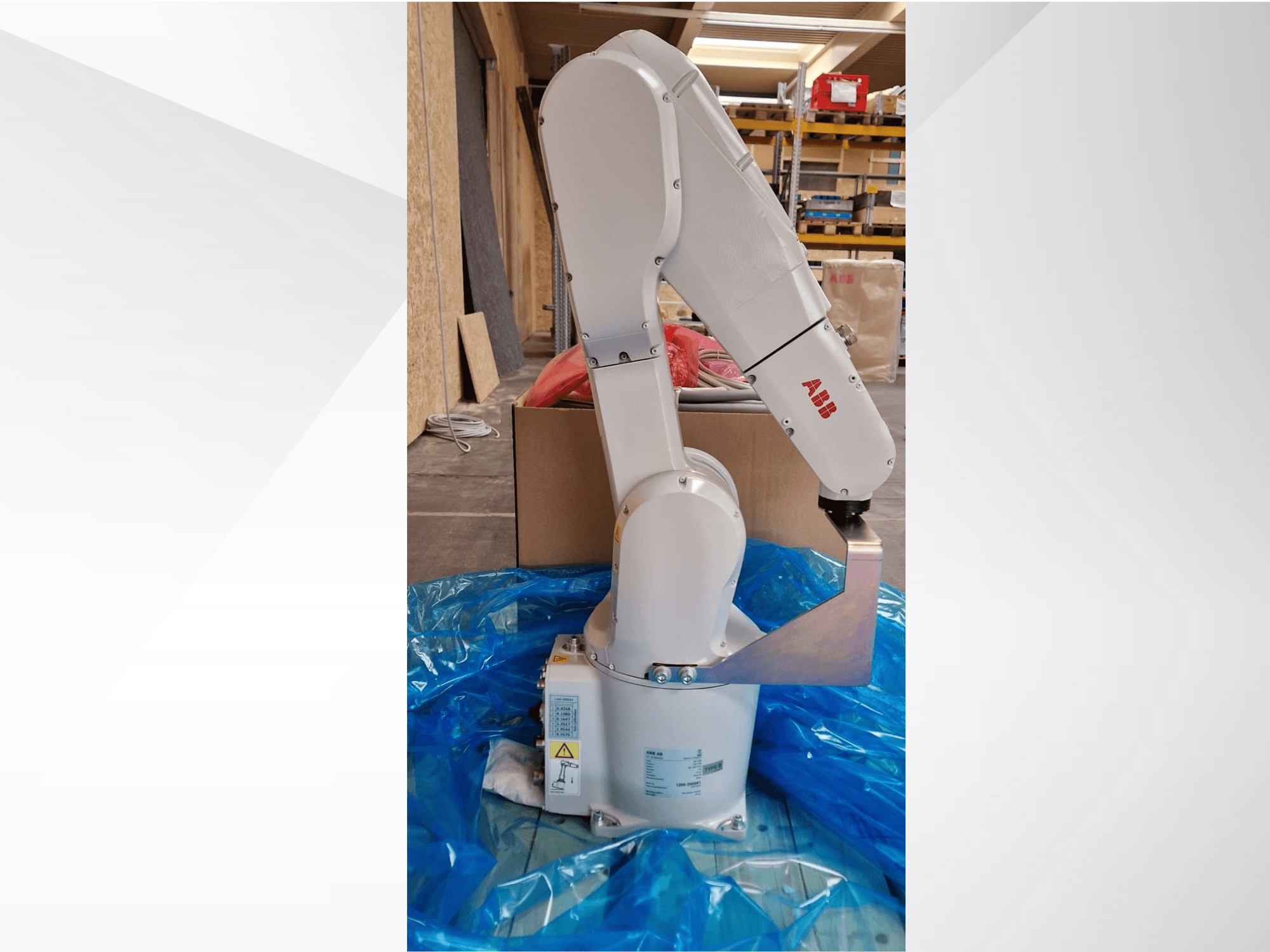 Modelo de braço robótico da ABB, vista lateral sobre lona azul, mostrando as juntas articuladas e a extremidade metálica, com etiquetas técnicas visíveis.