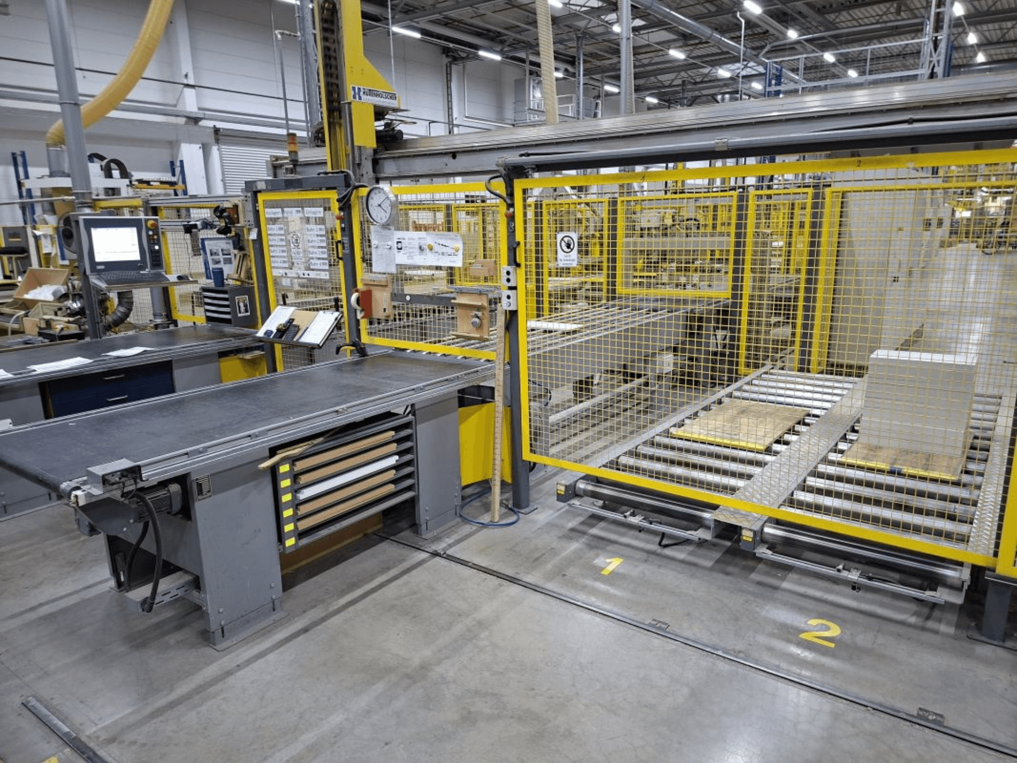 Espaço de trabalho industrial com um sistema de transporte e um painel de controlo; vista lateral mostrando a maquinaria e a vedação de segurança.