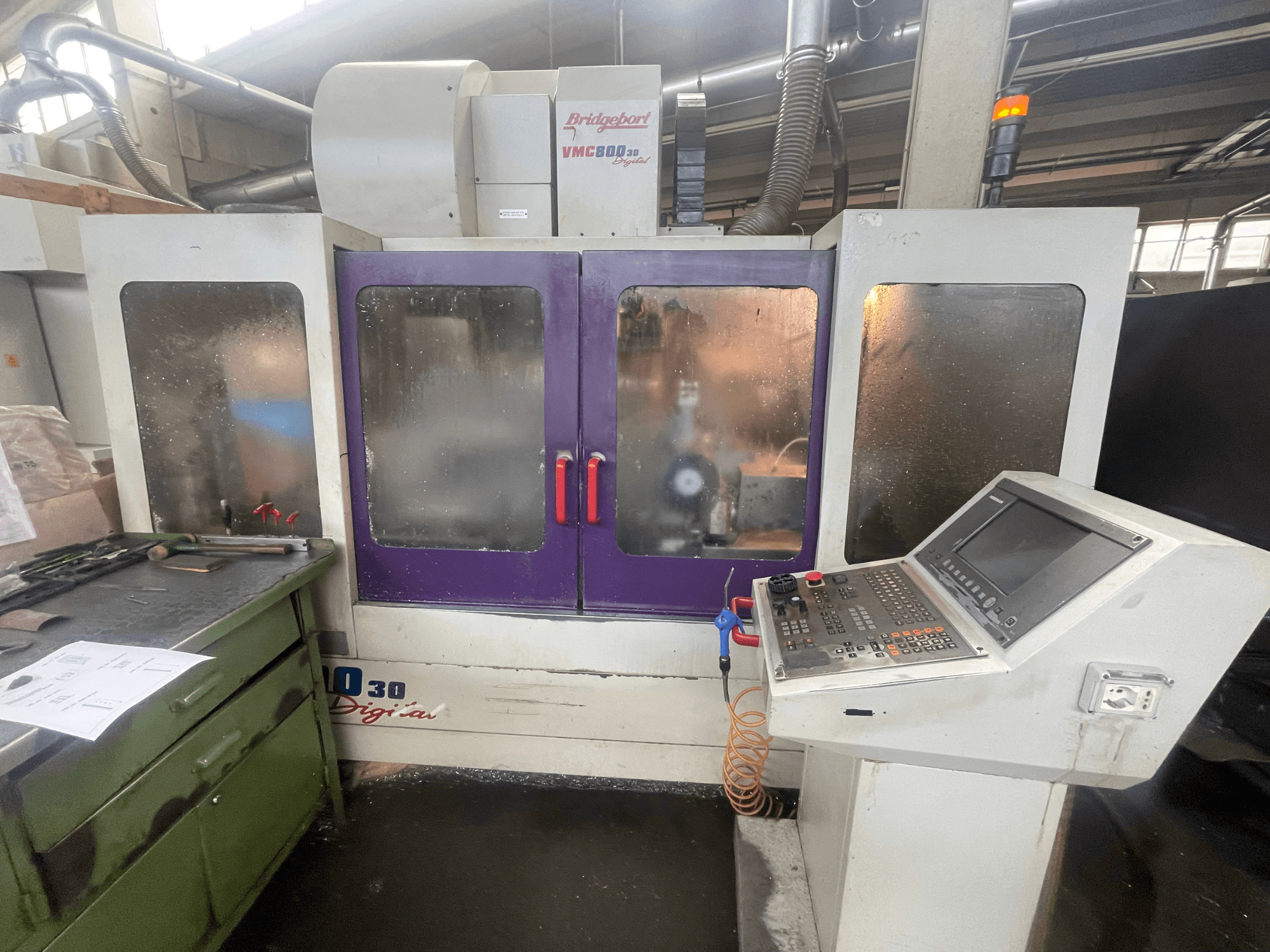 Centro de usinagem vertical Bridgeport VMC 800/30, vista frontal, mostrando o painel de controlo e as portas roxas, com ferramentas na superfície adjacente.