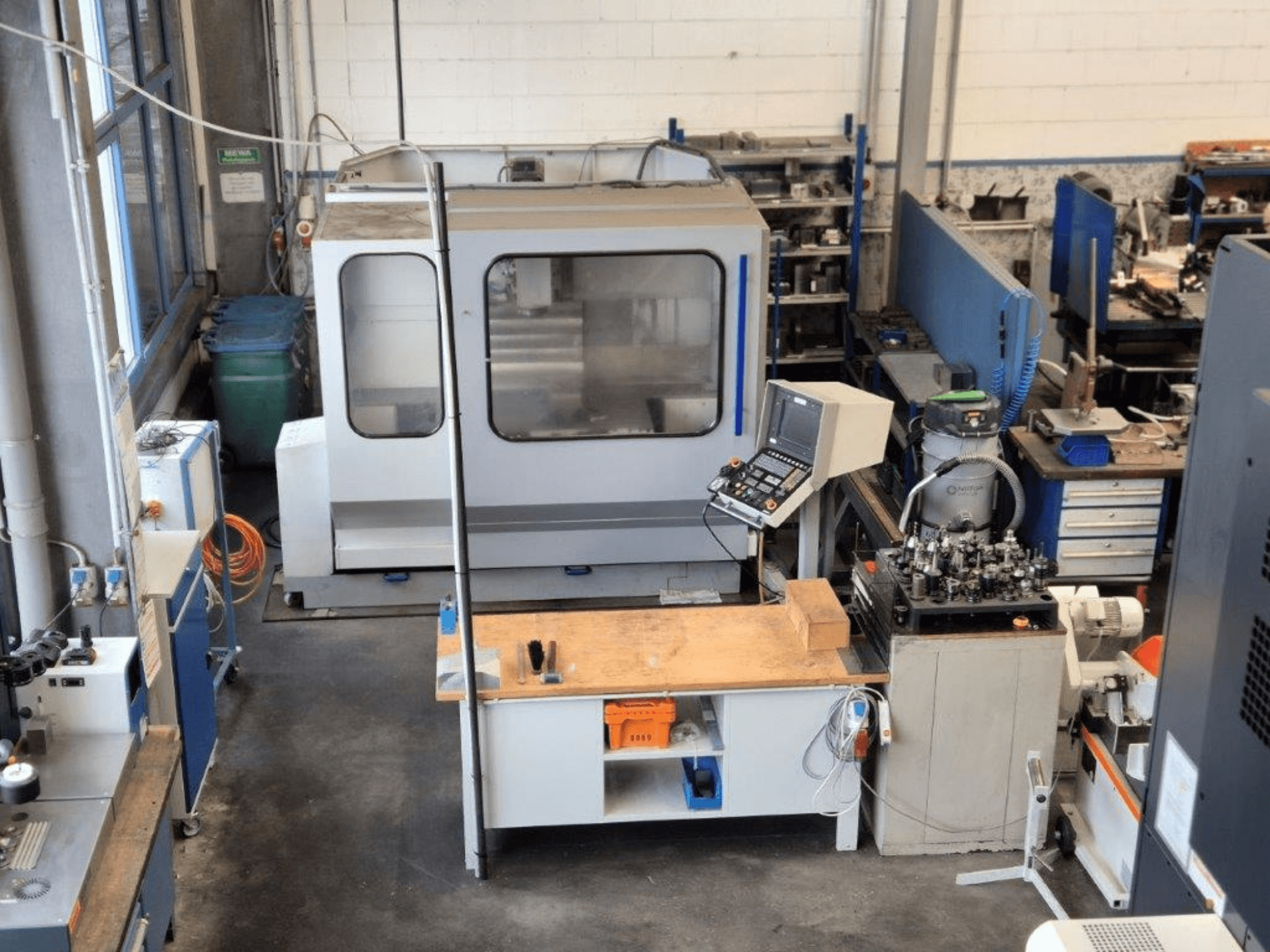 Fresadora CNC numa oficina, vista frontal, com painel de controlo, superfície de trabalho e ferramentas visíveis em segundo plano.