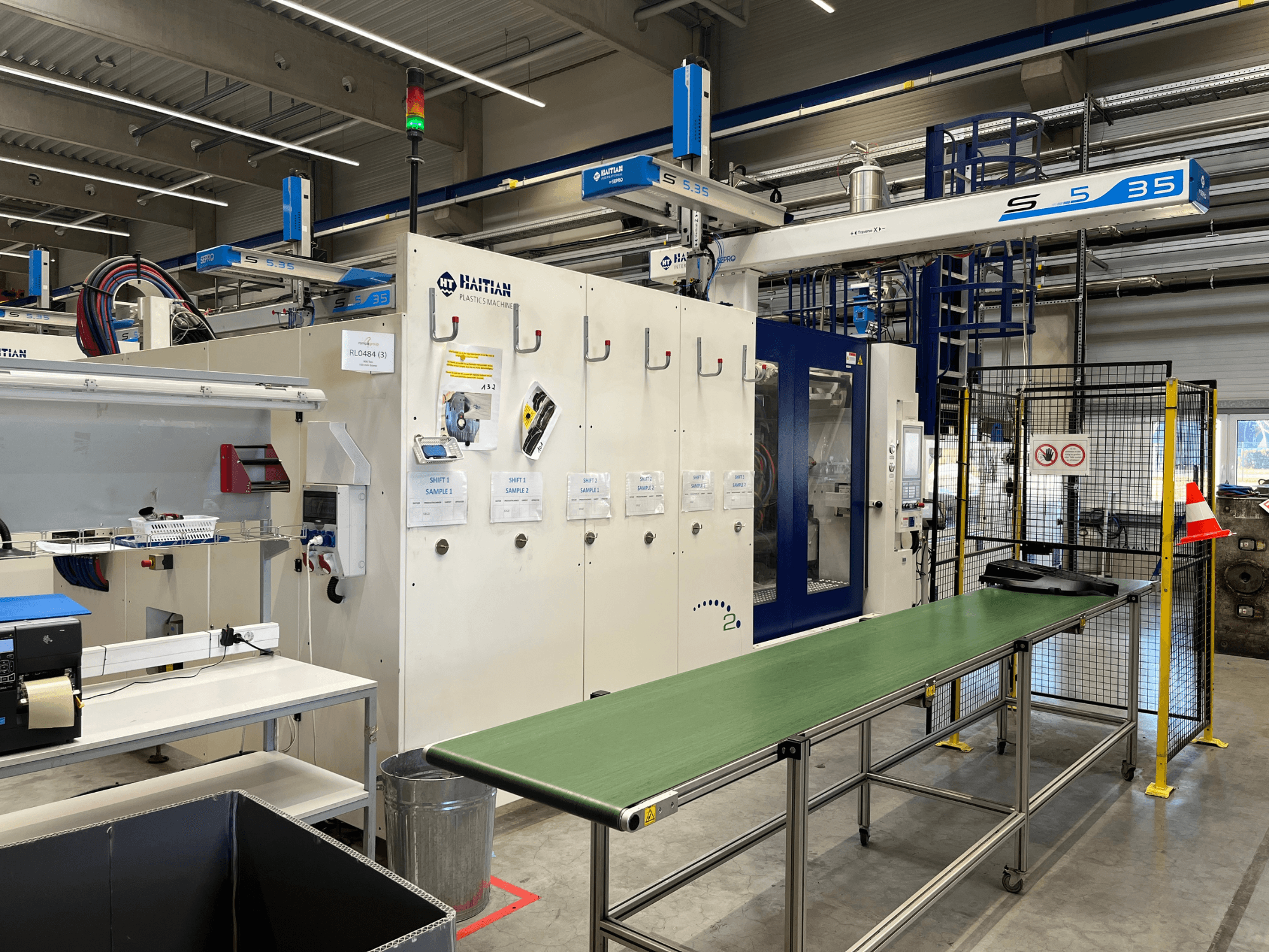 Máquina de plástico S5-35 haitiana numa oficina; mesa transportadora verde, barreiras de segurança e painel de controlo visíveis da vista frontal.