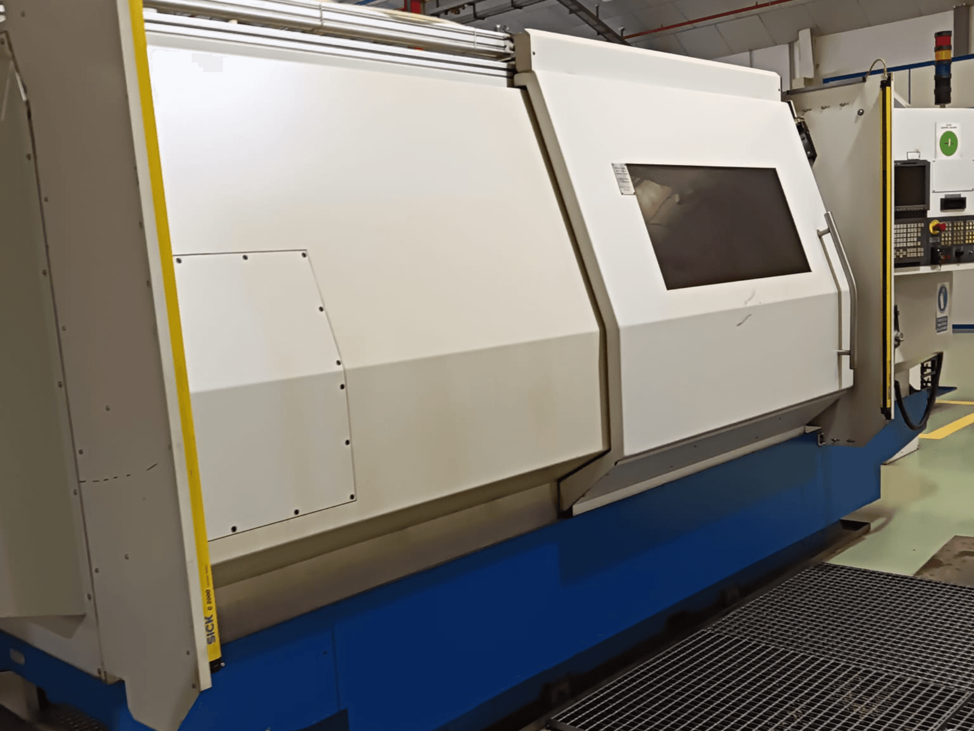 Máquina de torno industrial, vista lateral, com uma grande caixa branca e azul, painel de controlo digital e caraterísticas de segurança.