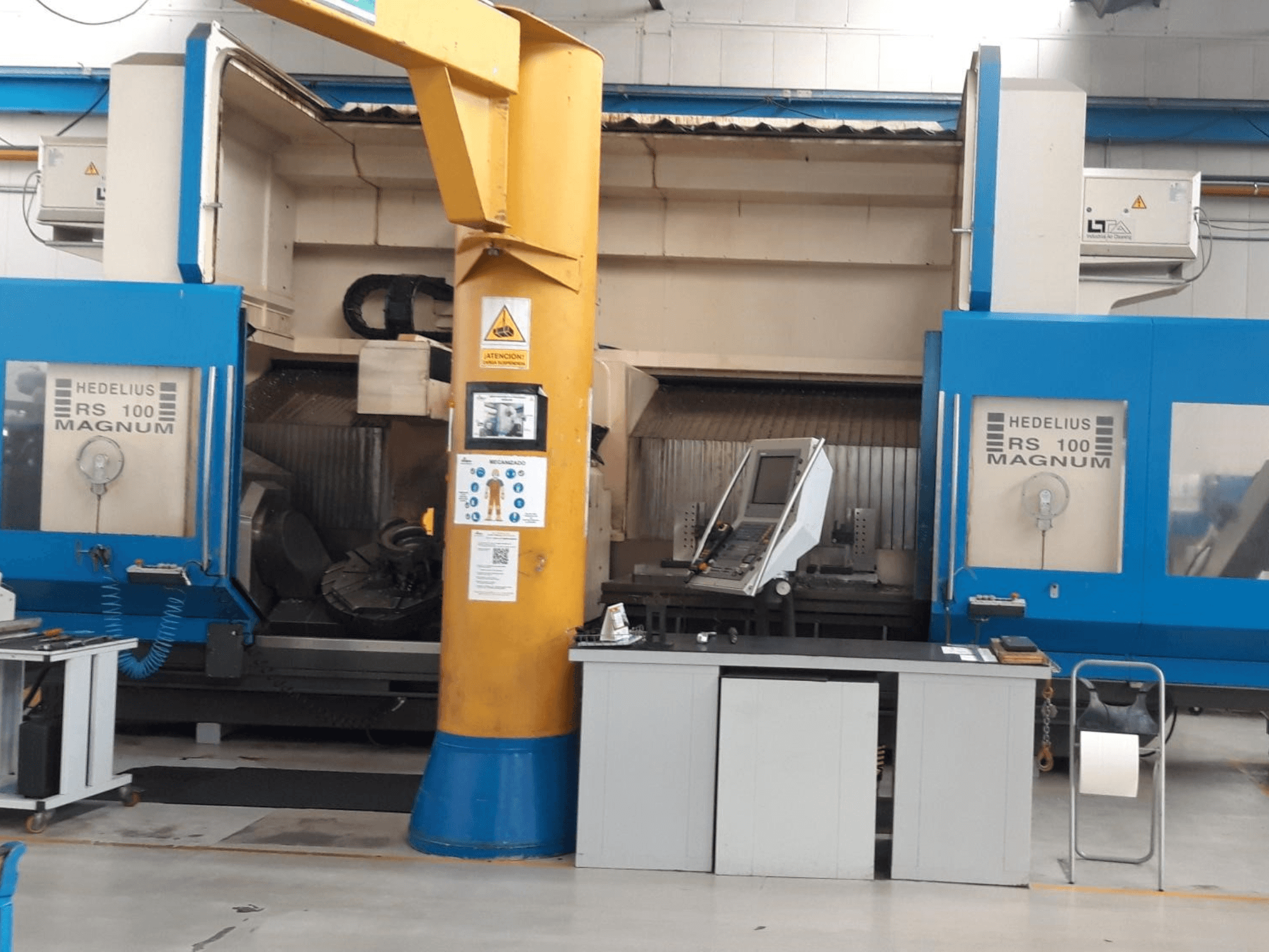 Máquina HEDALIUS RS 100 MAGNUM, vista frontal; apresenta painel de controlo e coluna de suporte amarela, inserida num espaço de trabalho industrial.