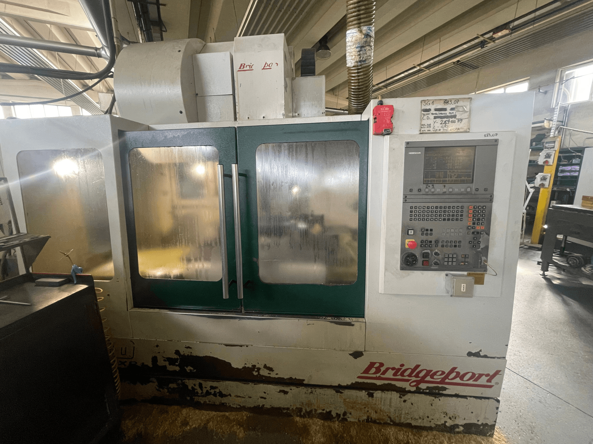 Centro de maquinagem vertical Bridgeport VMC 1000/30, vista frontal com painel de controlo, mostrando o exterior desgastado e as portas verdes.