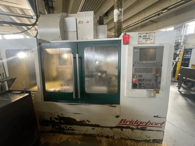 Centro de maquinagem vertical Bridgeport VMC 1000/30, vista frontal com painel de controlo, mostrando o exterior desgastado e as portas verdes.