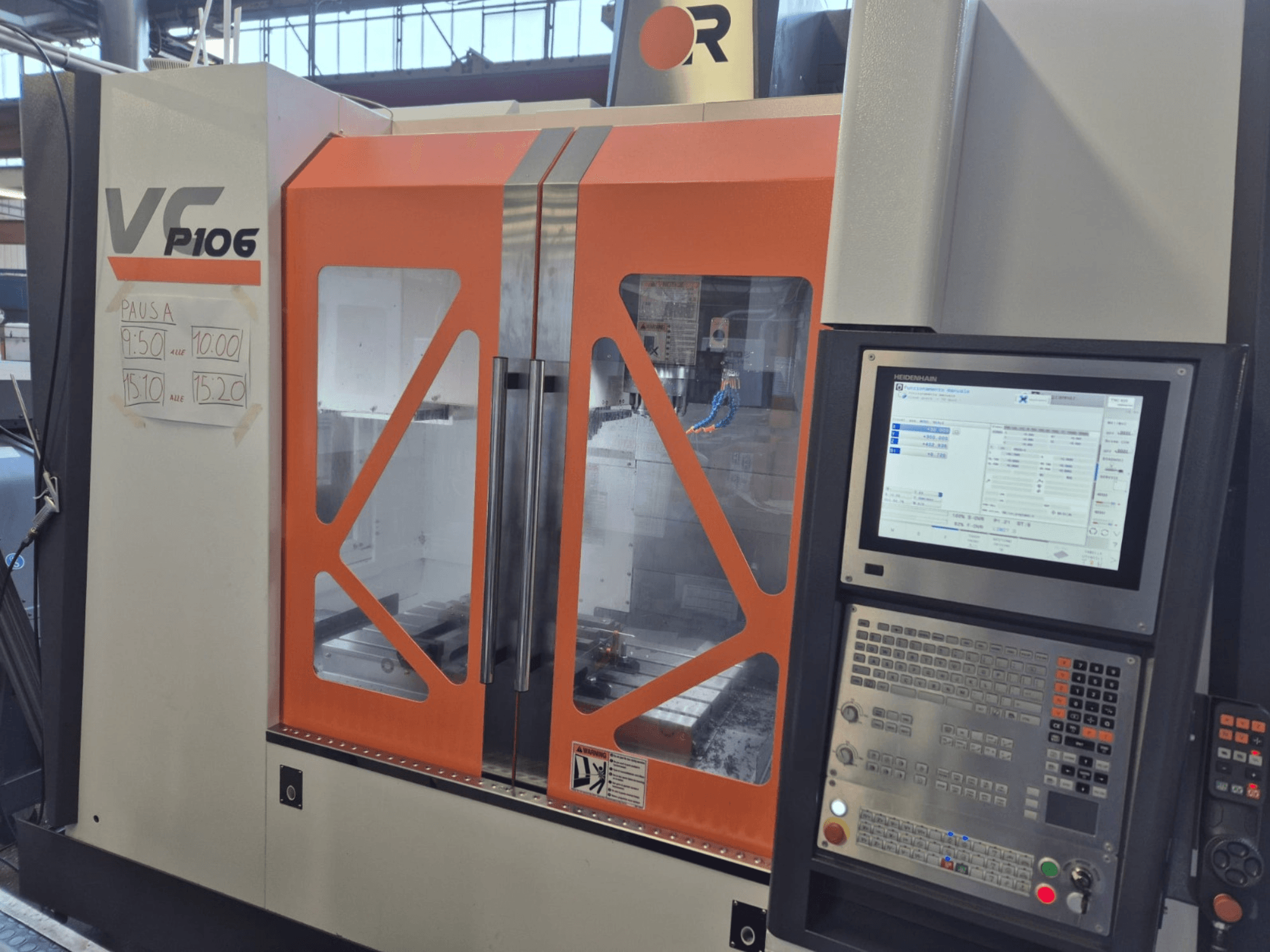 Texto alternativo: Máquina CNC VF P106 laranja e cinzenta, vista frontal, apresentando um painel de controlo e tempos de funcionamento programados em papel.
