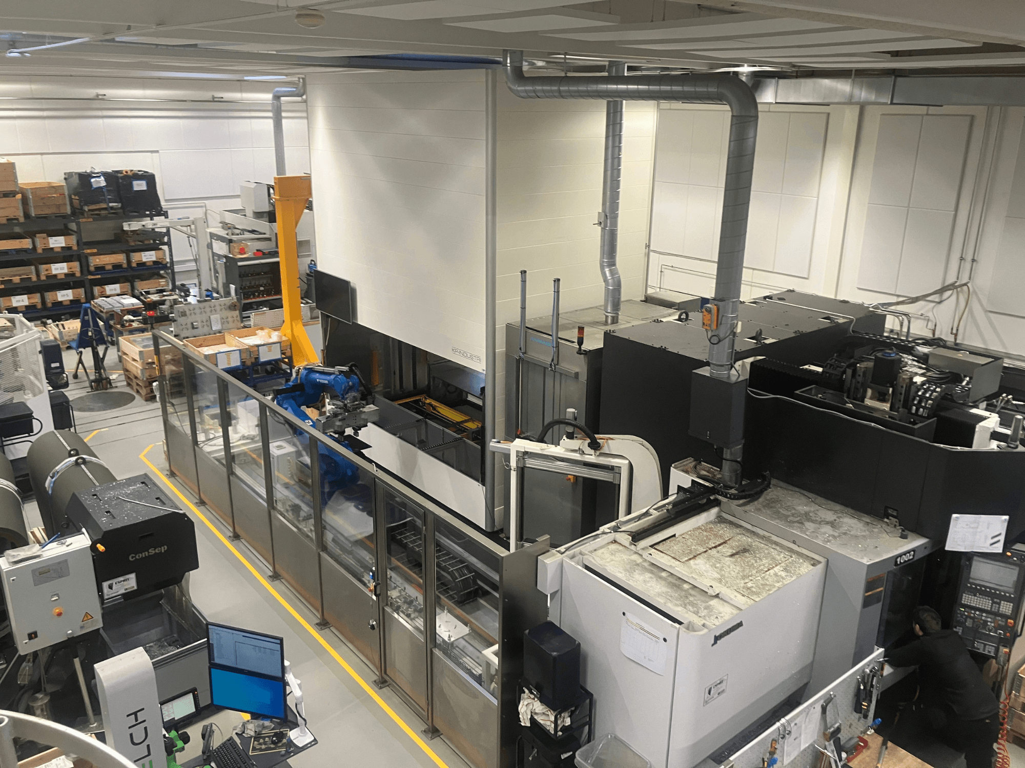 Sistema de automação Logimat da SSI SCHAFER num armazém, vista frontal; inclui braço robótico, prateleiras de armazenamento e máquinas.