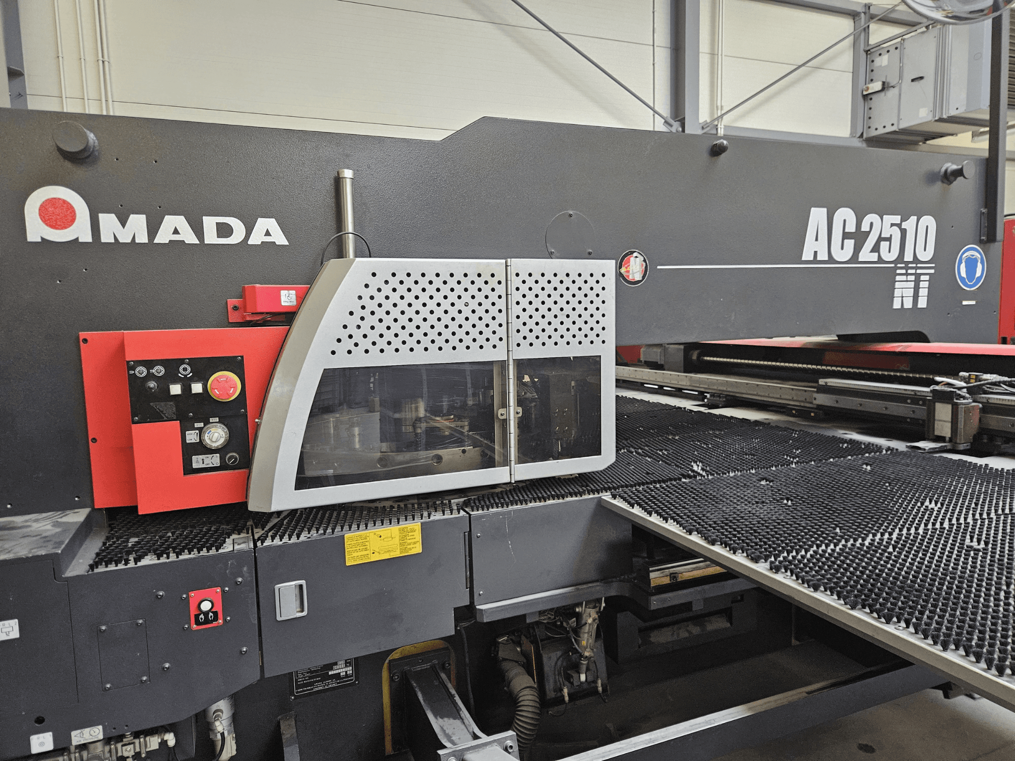 Máquina de corte a laser Amada AC2510 NT, vista frontal, com painel de controlo com botões e uma tampa de segurança.