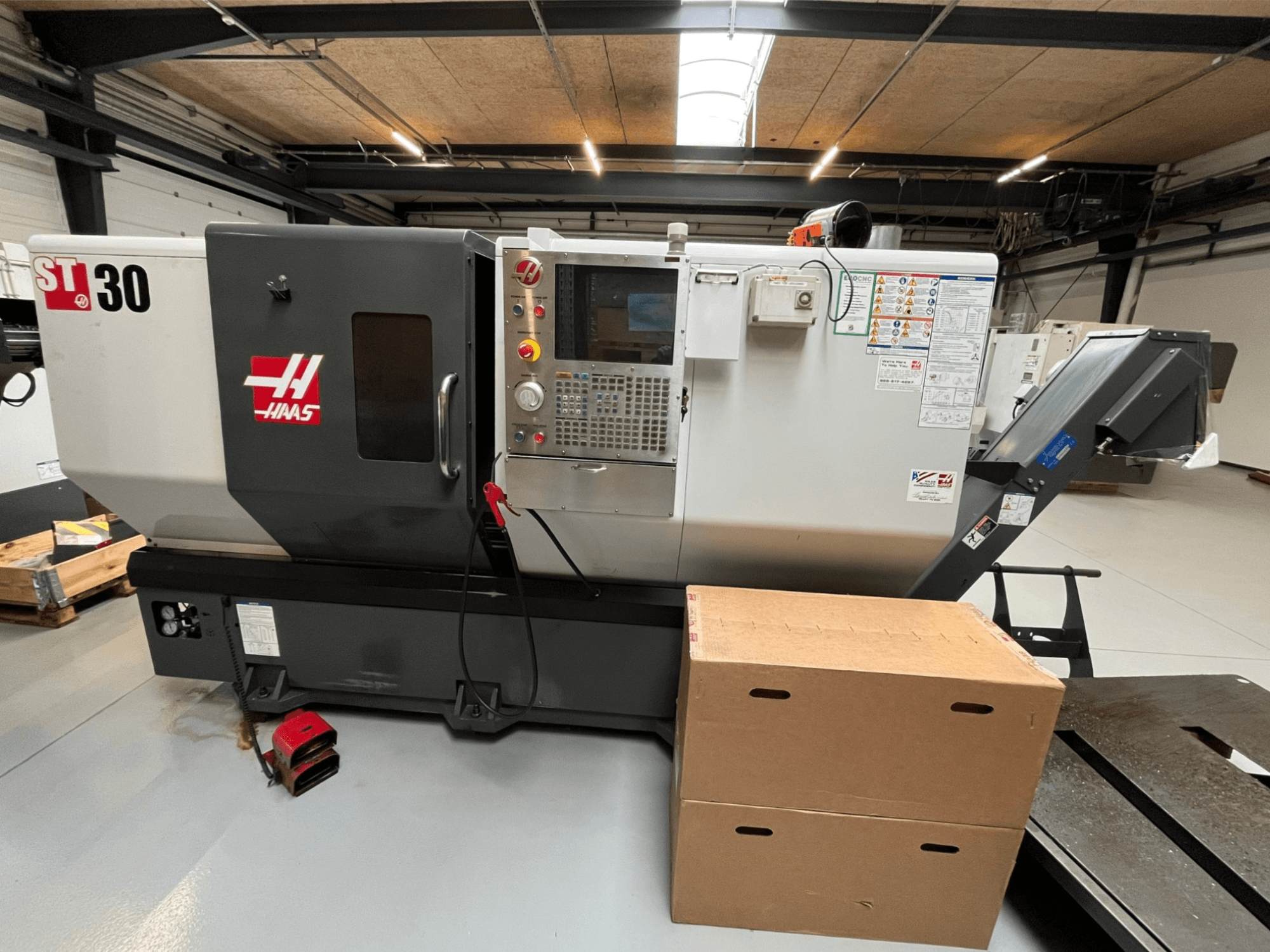 Torno CNC Haas ST-30, vista frontal, apresenta painel de controlo com botões, paragem de emergência e duas caixas de cartão empilhadas.