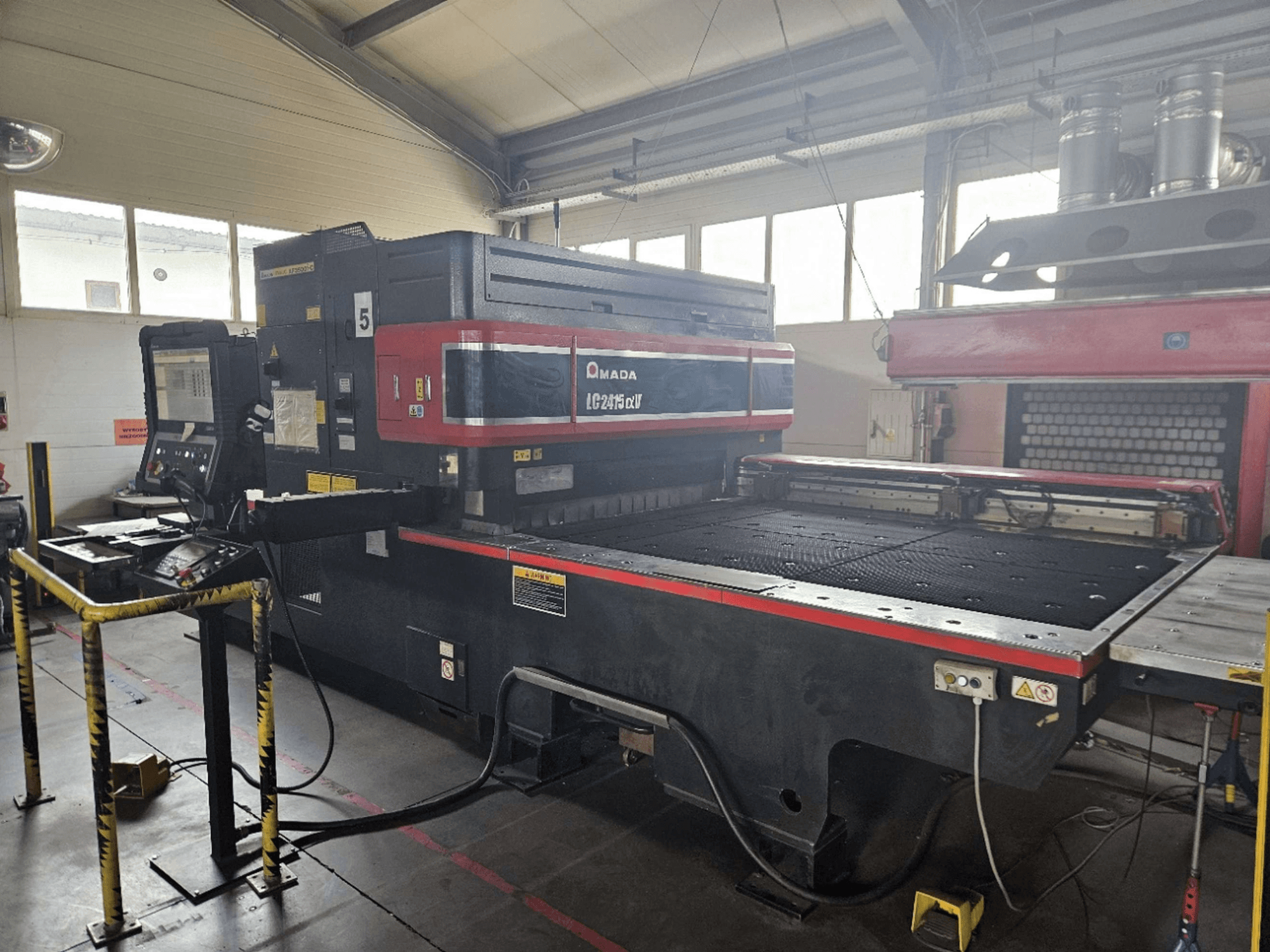 Máquina de corte a laser de CO₂ AMADA LC2415 Alpha 5, vista frontal que mostra o painel de controlo e a mesa de corte com barreiras de segurança.