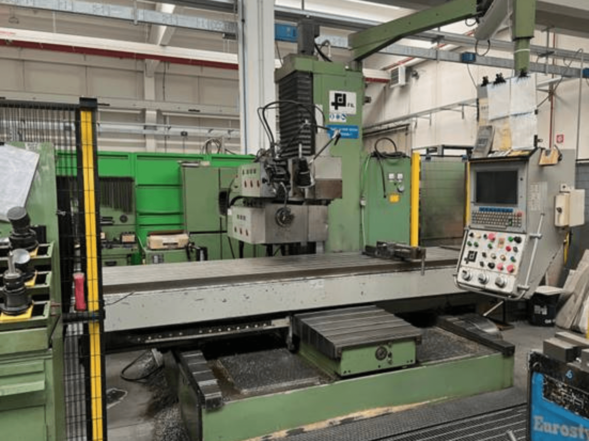 Fresadora industrial da JFL, vista frontal com painel de controlo, grande mesa plana e aparas de metal no chão.