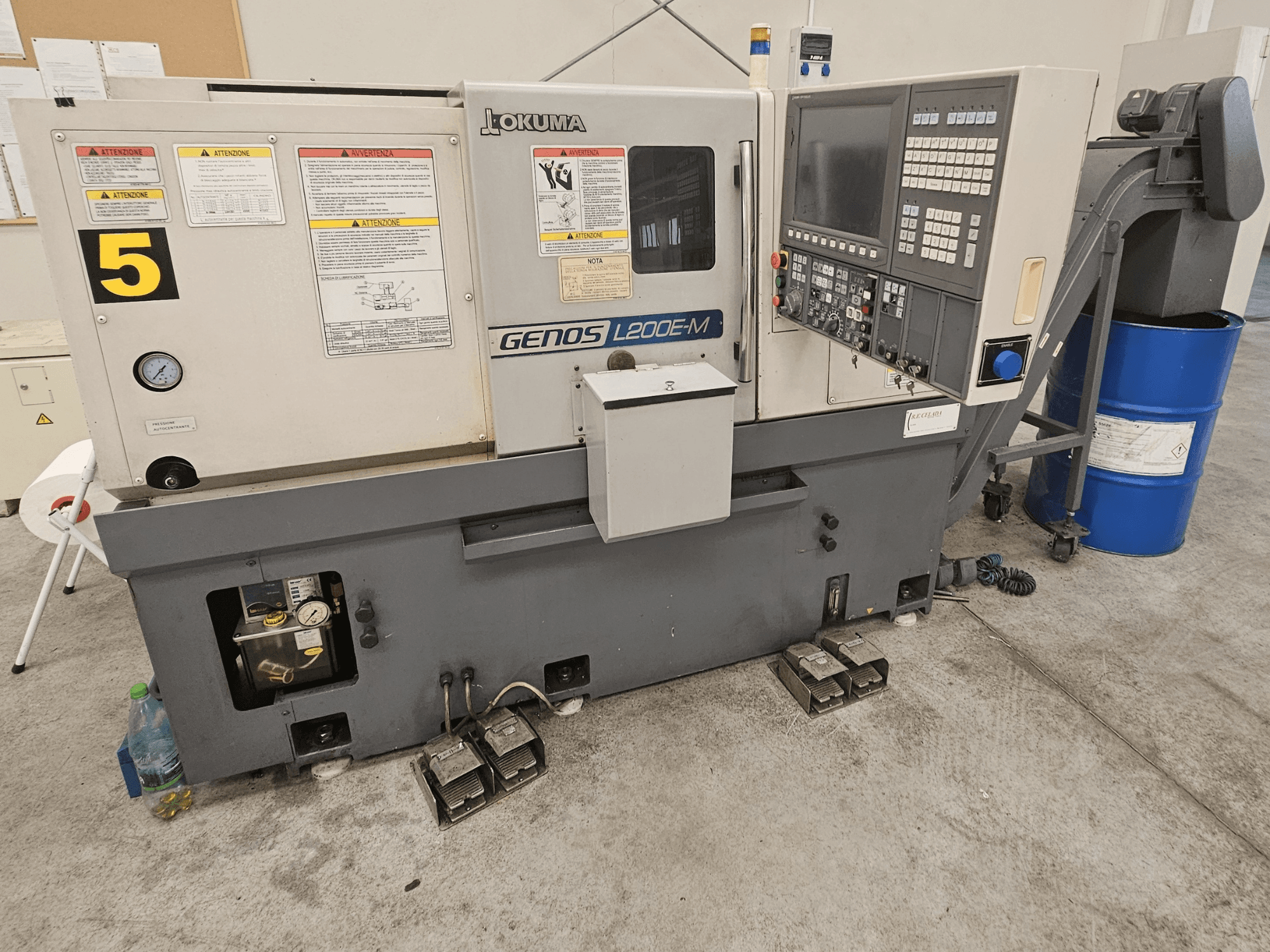 Vista frontal do torno horizontal Okuma GENOS L200E-M, com painel de controlo, etiquetas e tubo de refrigeração.