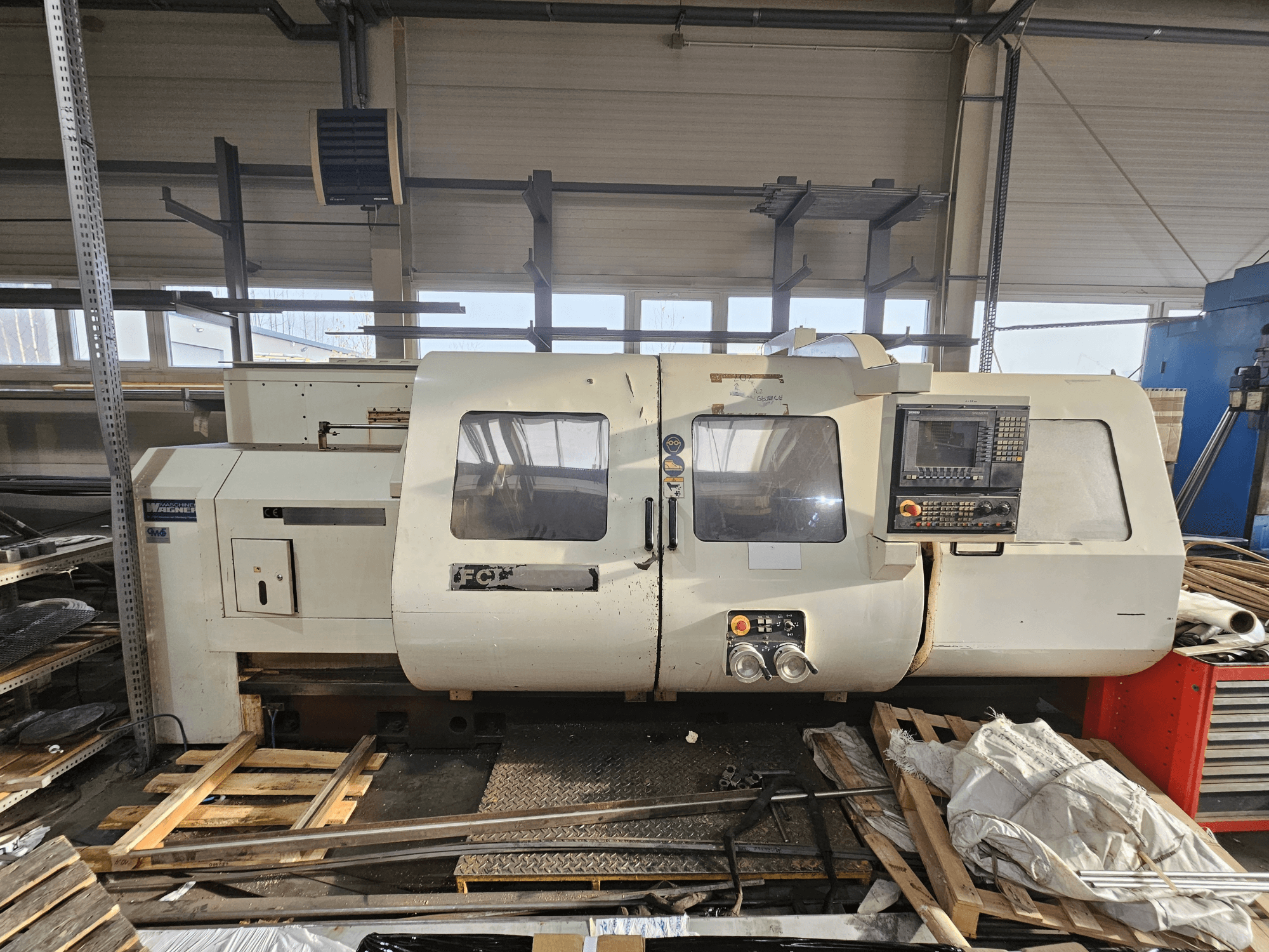 Torno CNC Wabeco, vista lateral, com painel de controlo, janelas e área de trabalho; máquinas sobre paletes de madeira em ambiente de oficina.