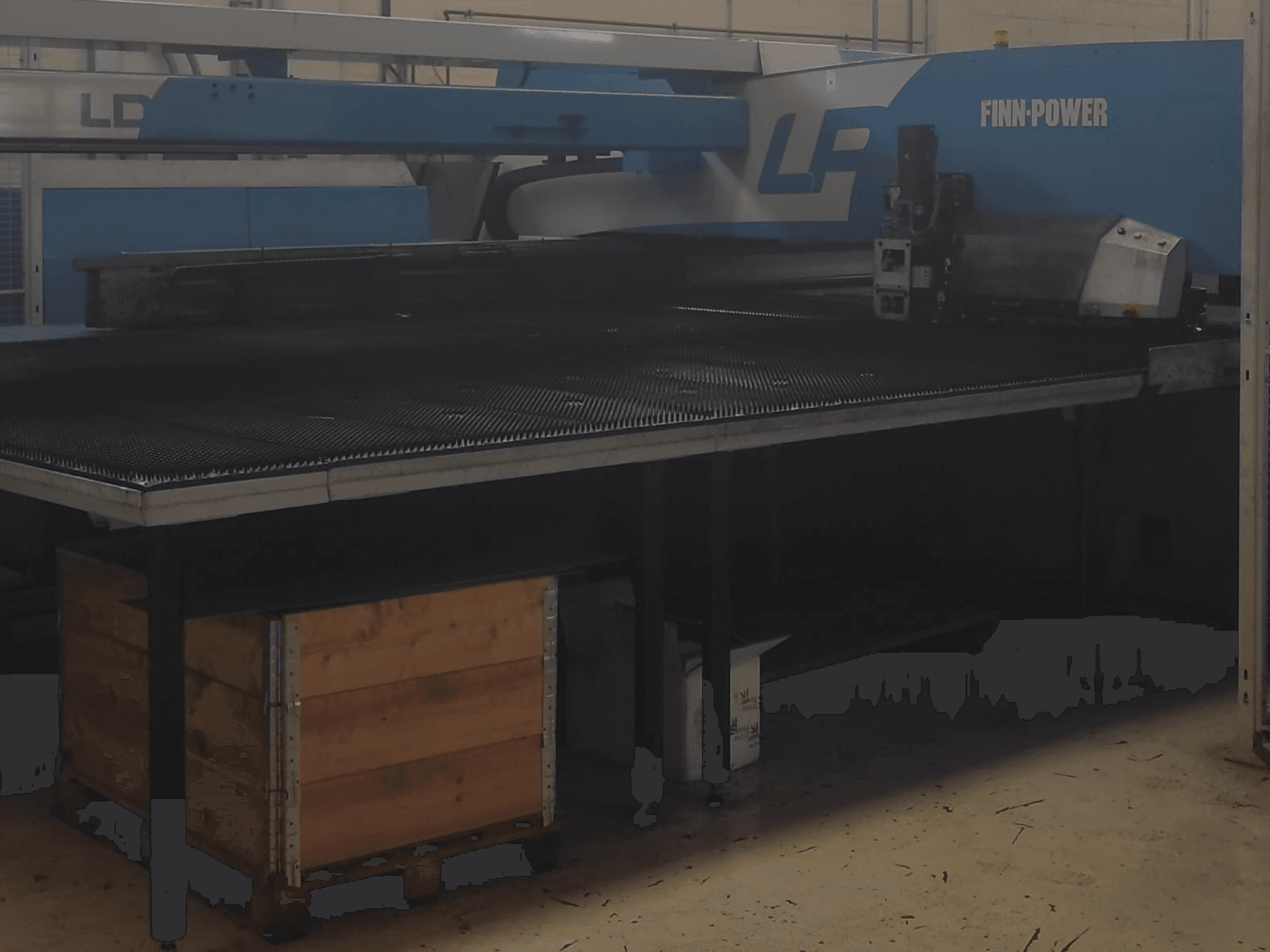 Máquina de corte Finn-Power LP numa fábrica, vista frontal com uma mesa de metal texturado e uma caixa de madeira por baixo.