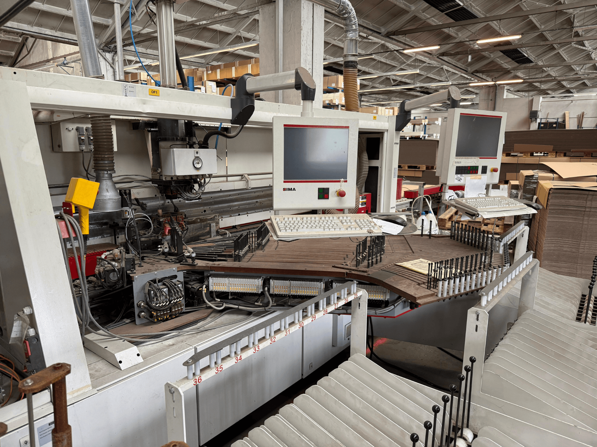 Máquina de precisão para trabalhar madeira IMA com dois monitores e painéis de controlo, vista frontal mostrando o transportador e a área de ferramentas.