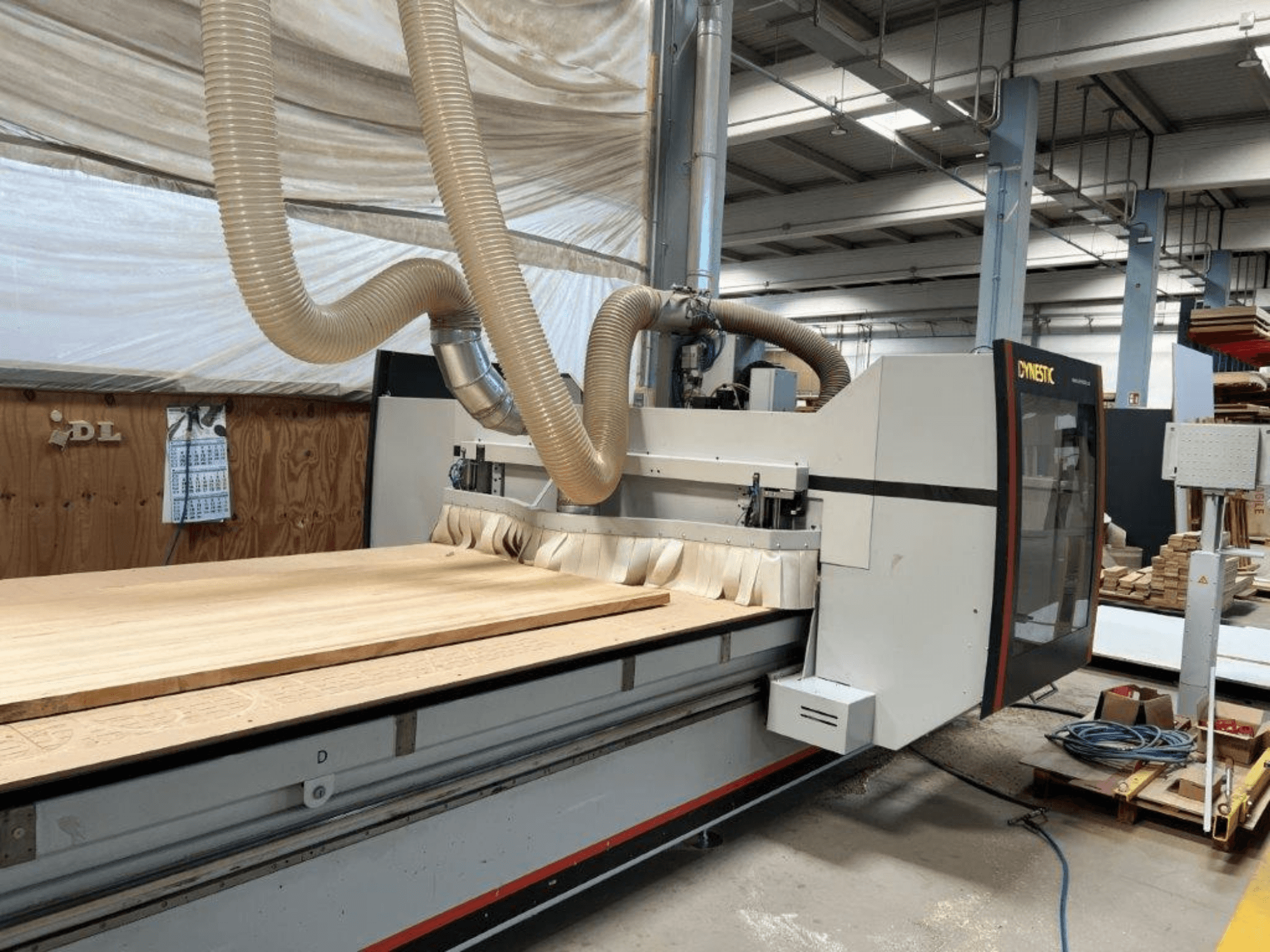 Máquina CNC da Dynestic, vista frontal, com uma grande peça de trabalho em madeira e mangueira de extração de poeiras numa oficina.