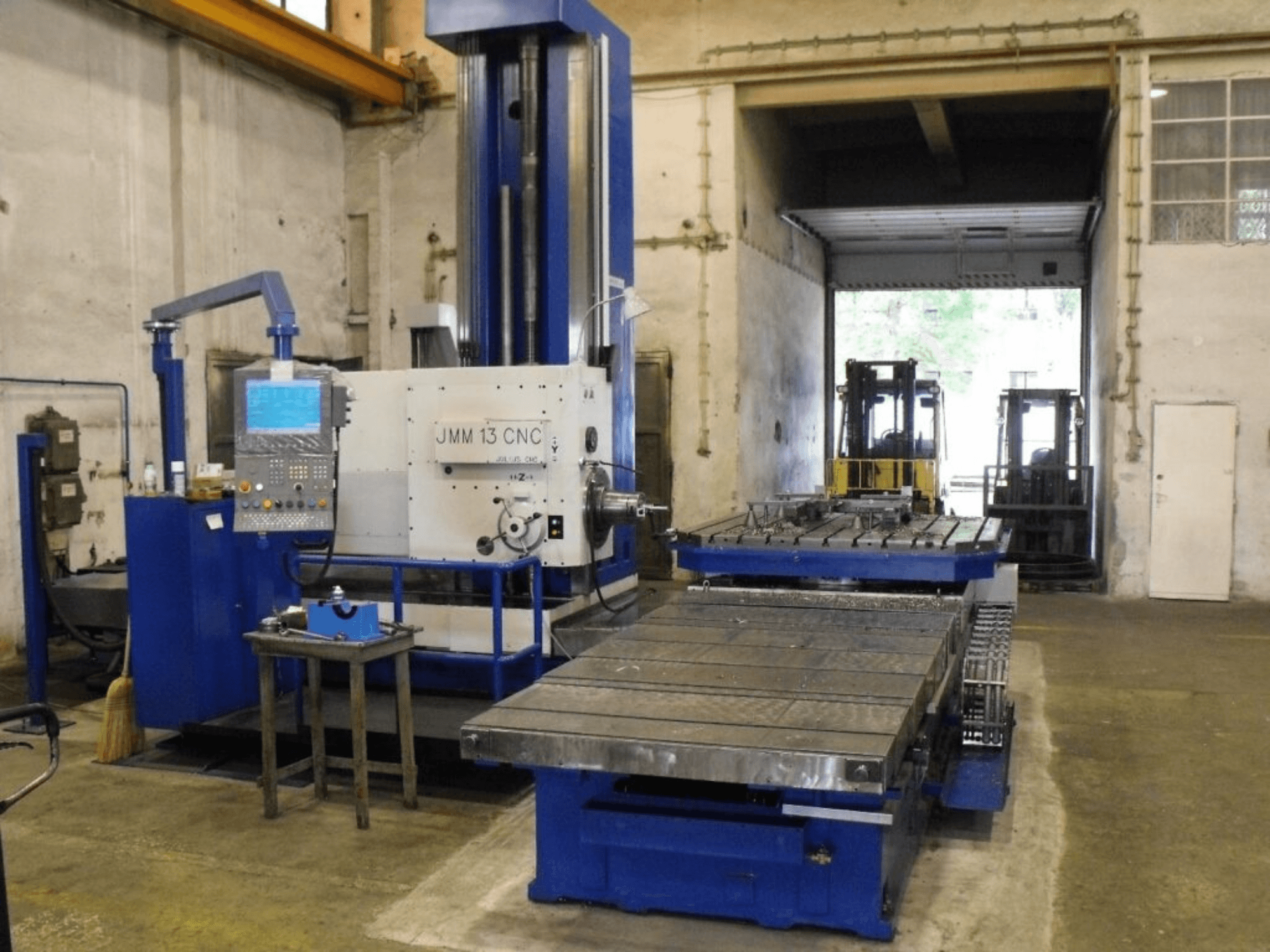 Fresadora CNC JMM 13 numa oficina, vista frontal, com um painel de controlo, uma grande mesa de trabalho e um pano de fundo industrial.