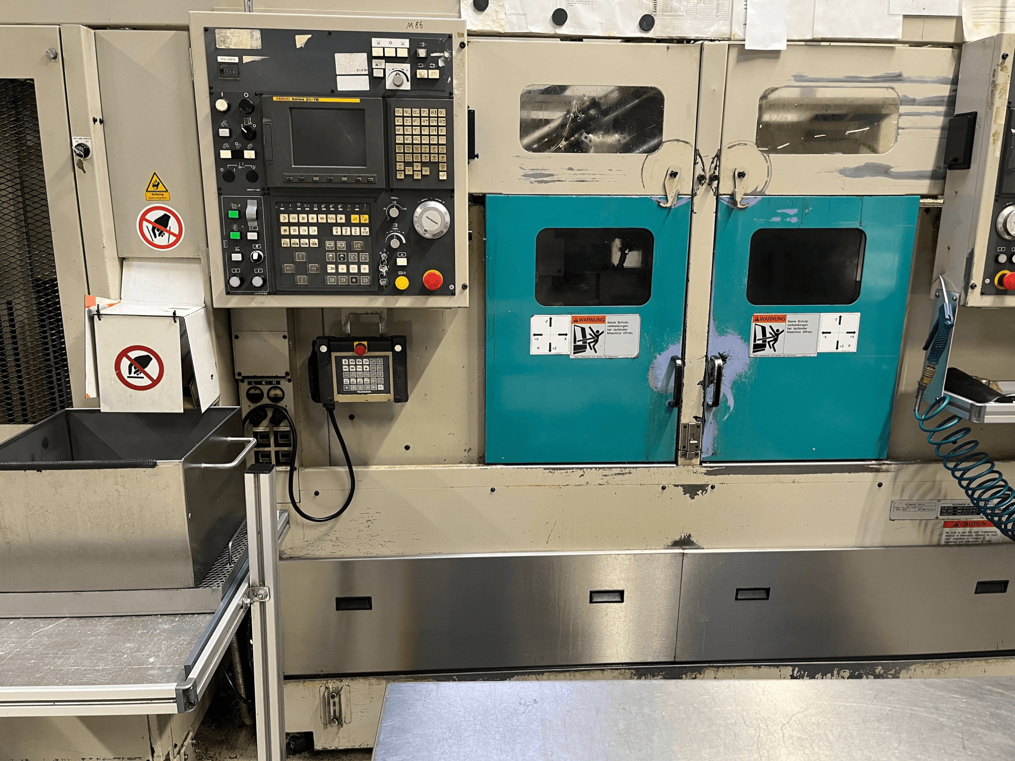 Painel de controlo de uma máquina de fabrico com portas em azul-petróleo, avisos de segurança e um teclado; vista frontal, acabamento em aço inoxidável.