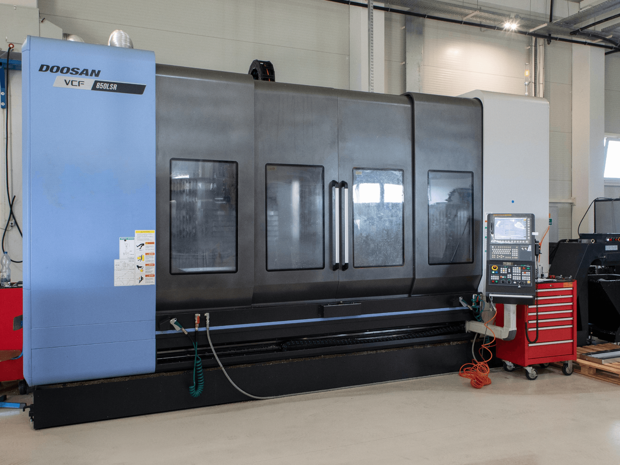 Centro de fresagem e torneamento DOOSAN VCF850, vista frontal; apresenta janelas duplas de grandes dimensões, painel de controlo e compartimento para ferramentas. Ideal para usinagem.
