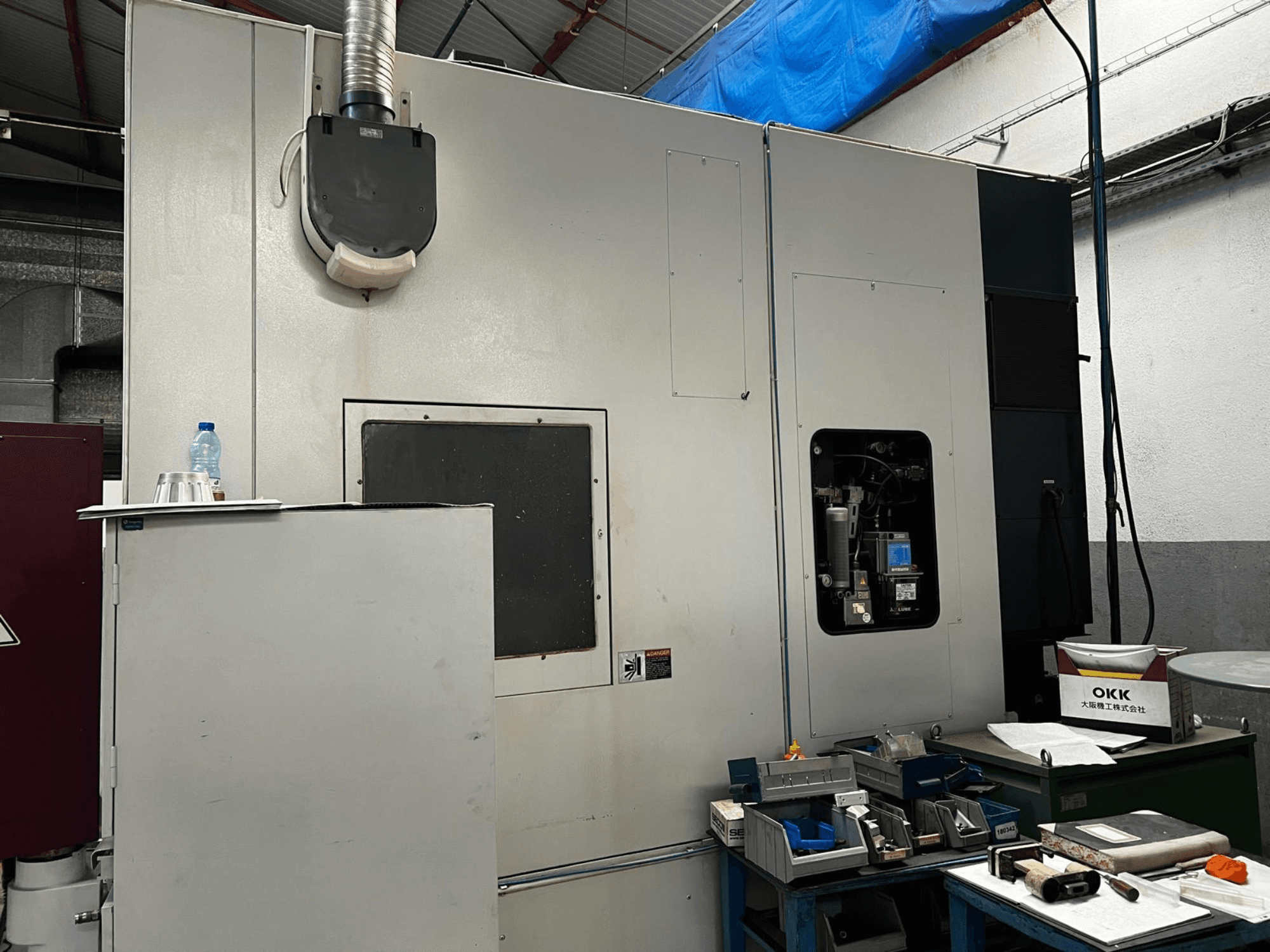 Centro de usinagem vertical OKK VP600 numa oficina, vista lateral que mostra os detalhes da máquina e as ferramentas numa mesa próxima.