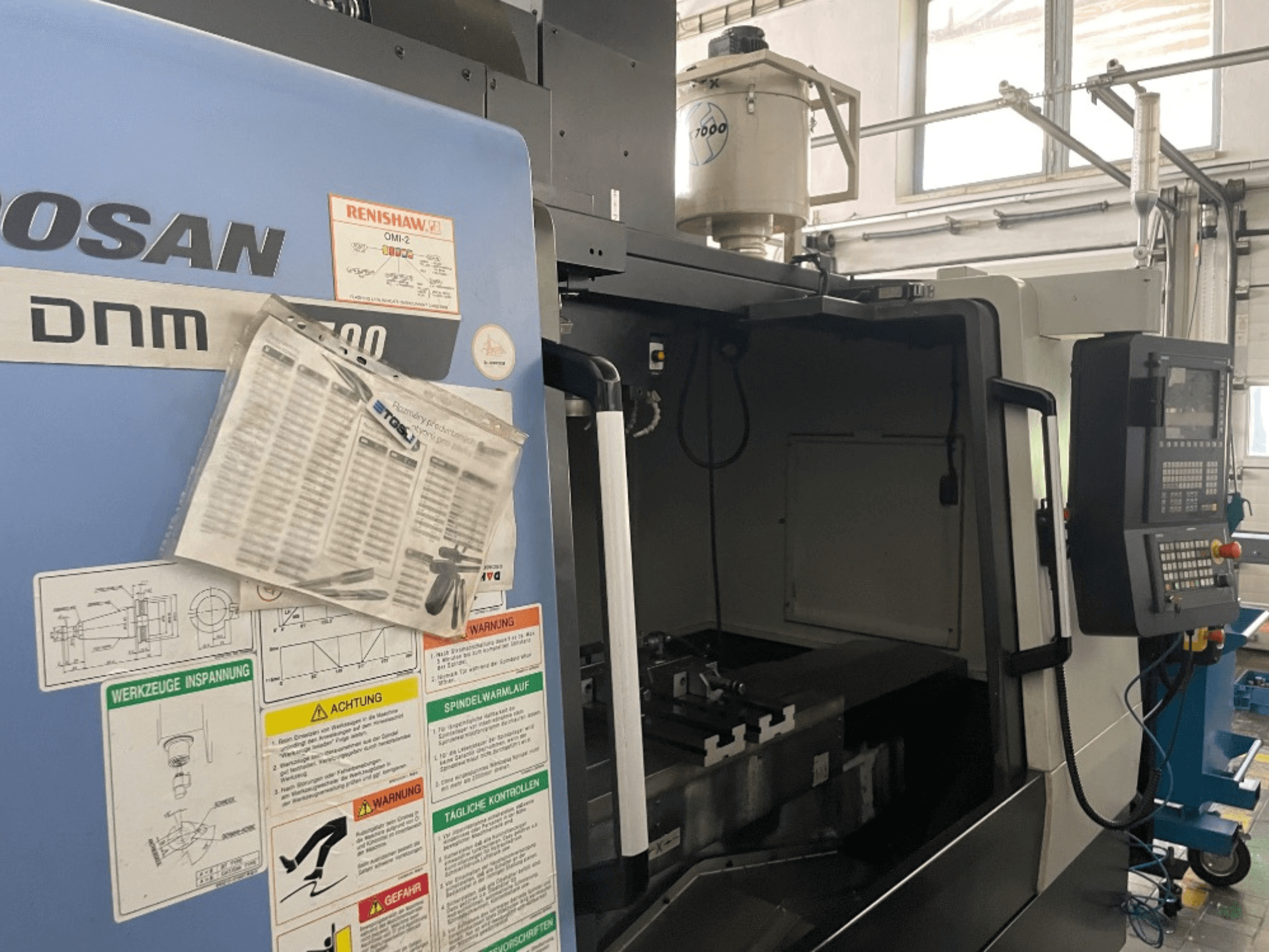 Centro de maquinação vertical DOOSAN DNM 5700, vista frontal, mostra o painel de controlo, etiquetas de segurança e detalhes da máquina.