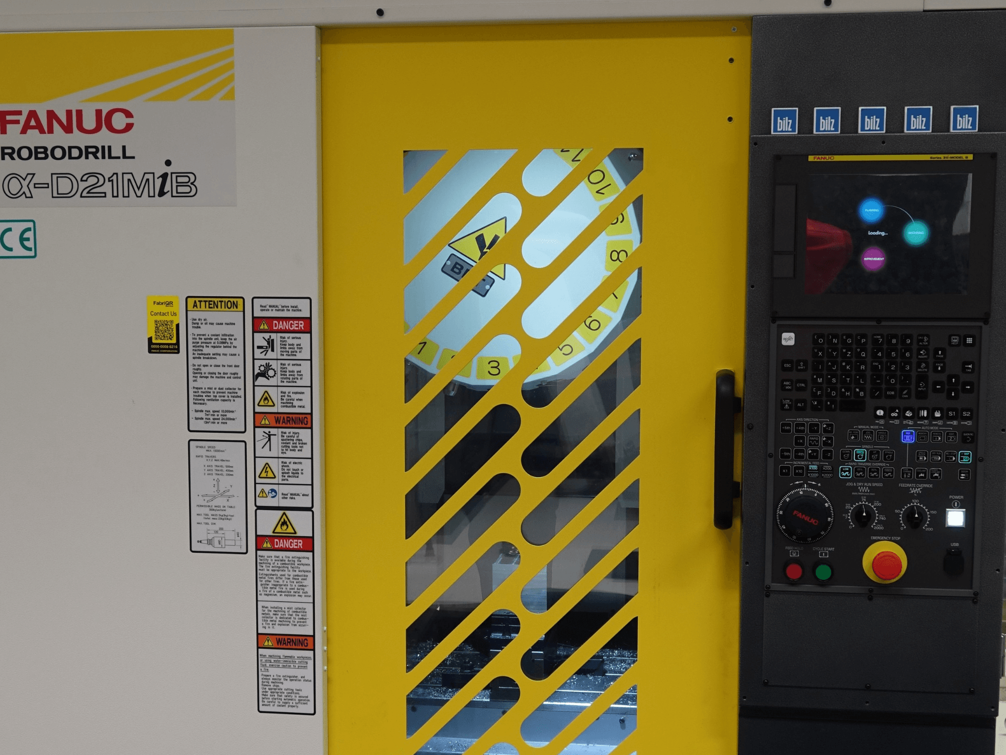 Vista frontal do Centro de Maquinação Vertical FANUC α-D21MiB, mostrando as advertências de segurança, o painel de controlo e a porta de segurança amarela.