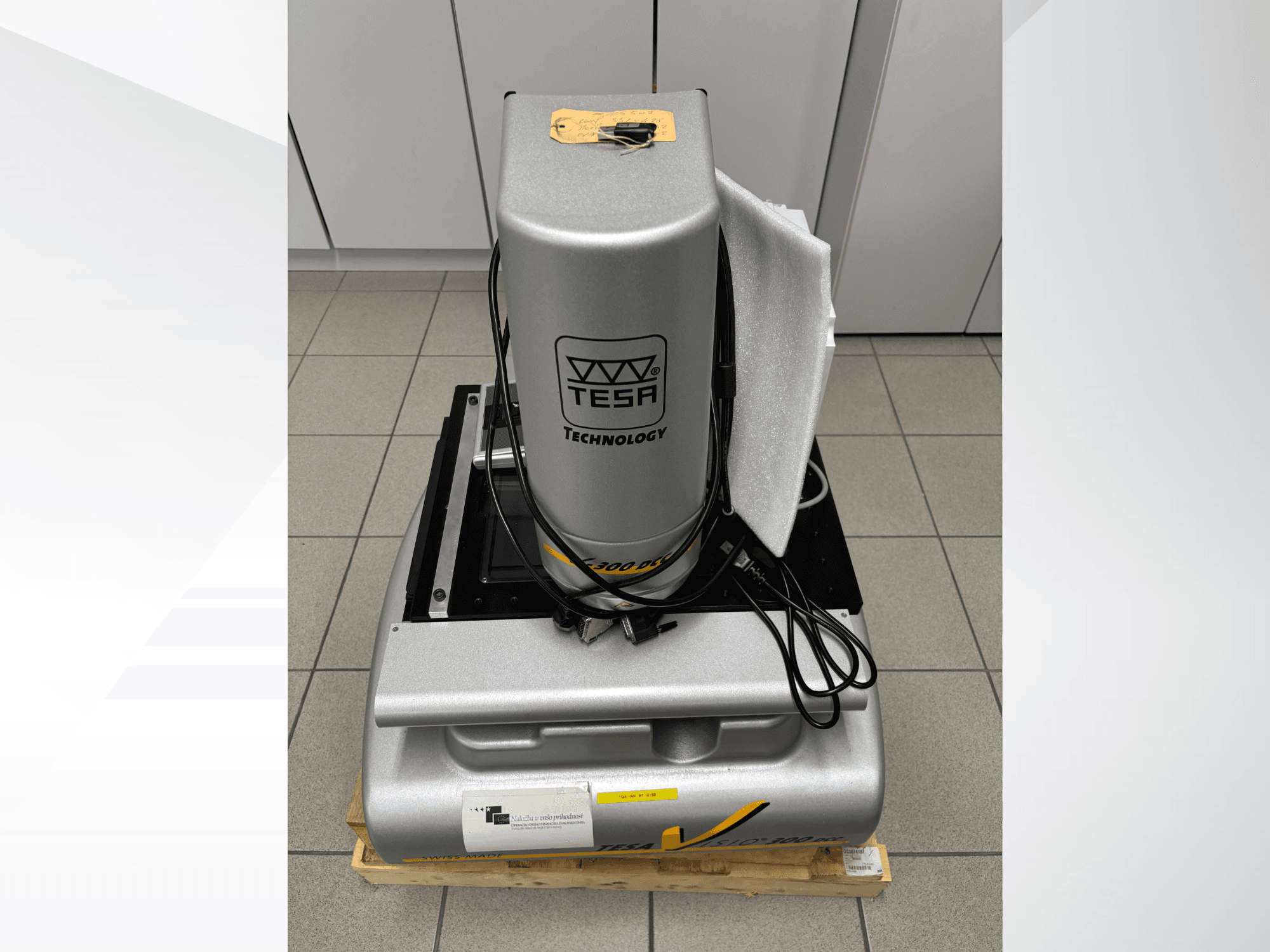 Máquina de medição ótica TESA Visio 300 DCC em vista frontal com cabos, numa palete de madeira, e embalagem de proteção visível.