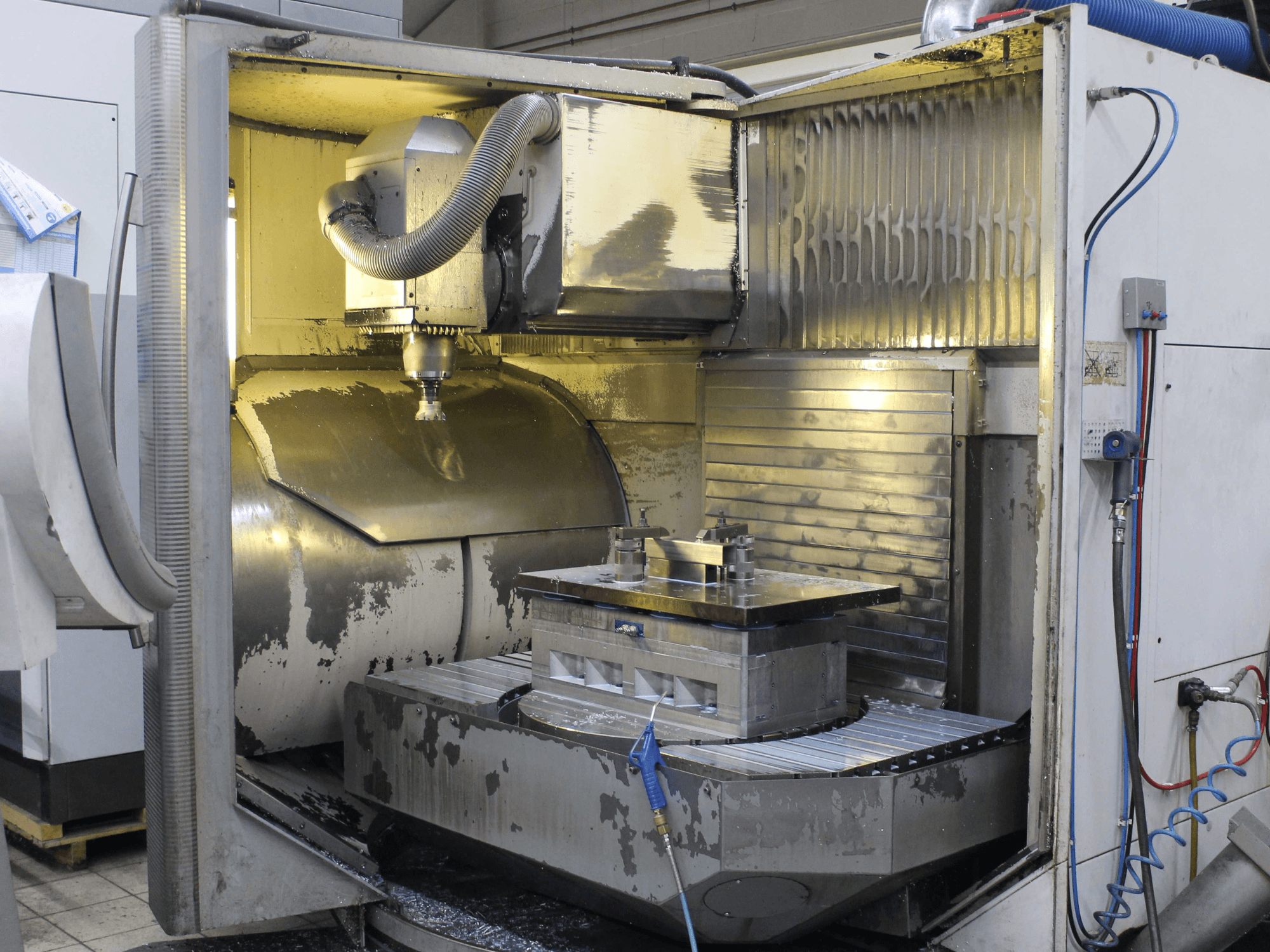 Interior de uma fresadora CNC, vista lateral, mostrando a peça de trabalho e a ferramenta de corte com aparas de metal e resíduos de líquido de refrigeração.