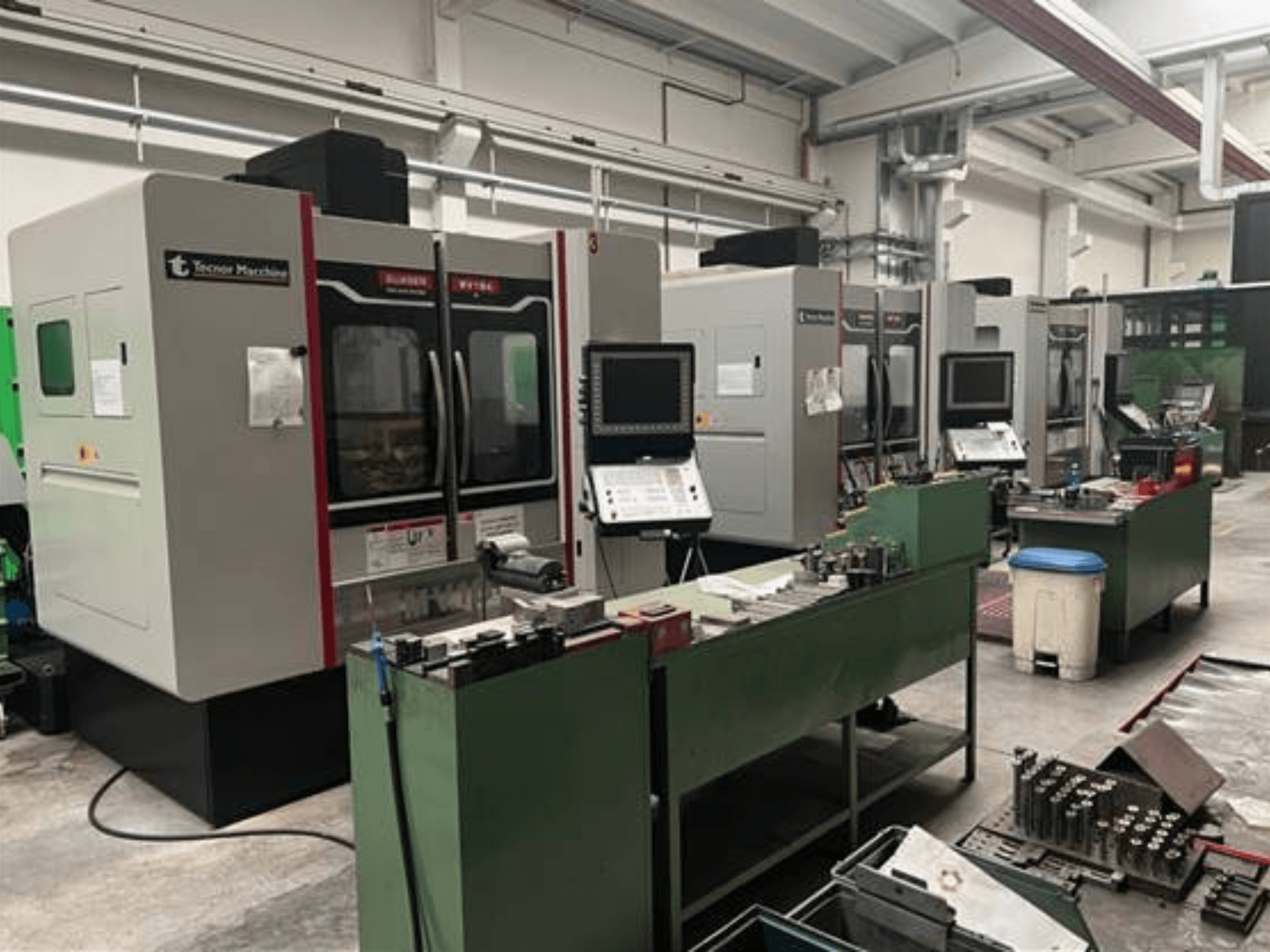 Quatro máquinas CNC da Tecnoer Marchine numa oficina, vista frontal, com ferramentas e peças em bancadas de trabalho verdes.