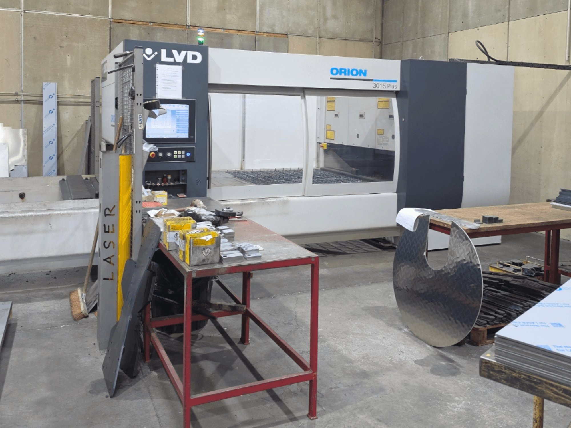Máquina de corte a laser de CO₂ LVD Orion 3015 Plus, vista frontal com painel de controlo; rodeada por peças metálicas sobre uma mesa de trabalho.
