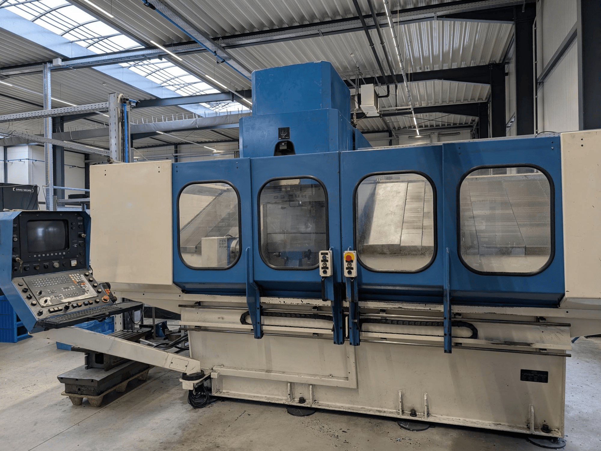 Máquina CNC industrial azul com painel de controlo, vista frontal. Grandes janelas de vidro, componentes da máquina e botões visíveis.