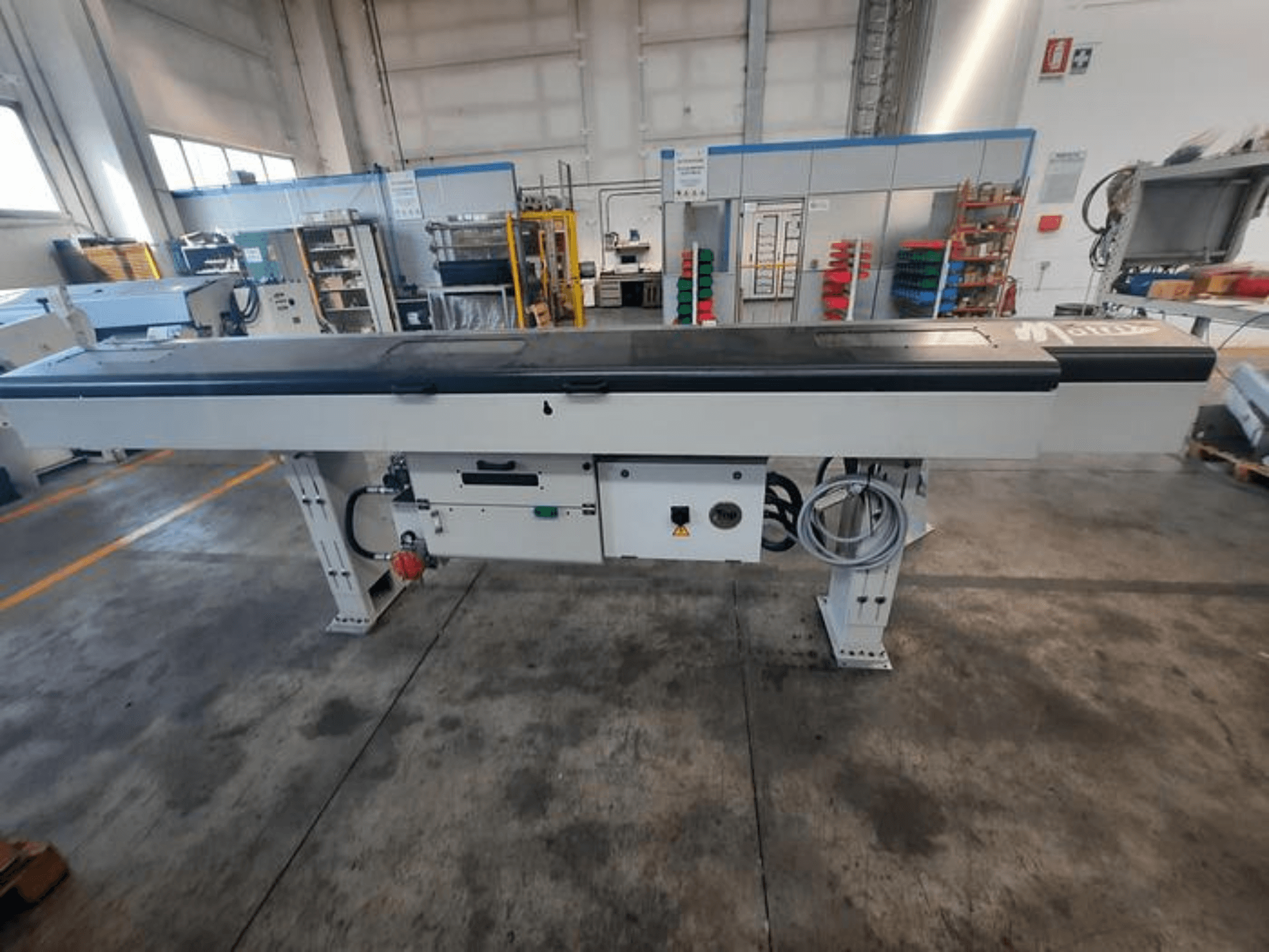 Mesa transportadora metálica com tampo preto liso, vista lateral, com botões e cabos de controlo; logótipo da marca parcialmente visível.