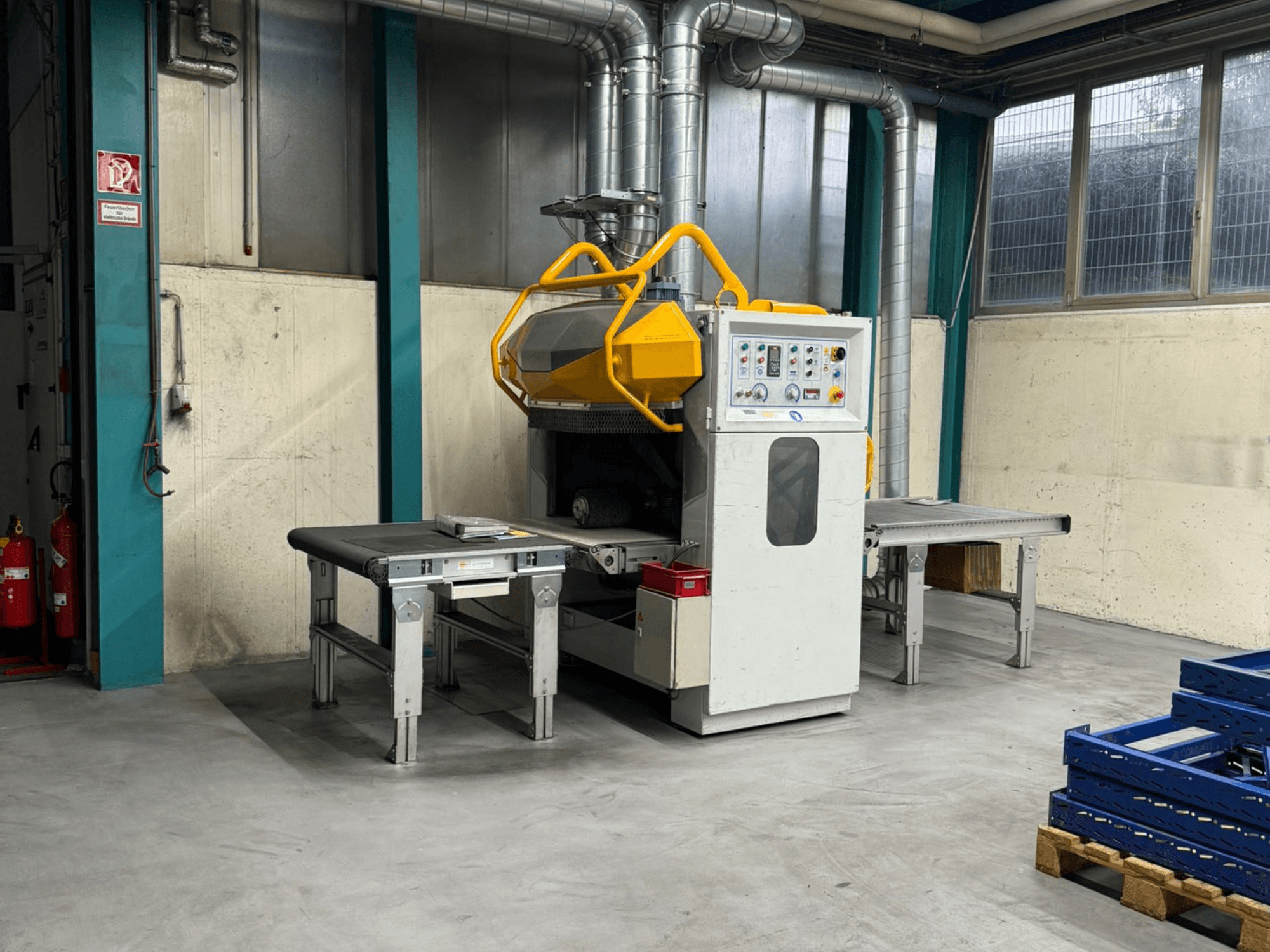 Máquina de rebarbação Fladder AUT-1000 VAC numa fábrica, com painel de controlo, proteção de segurança amarela e mesas de trabalho adjacentes.