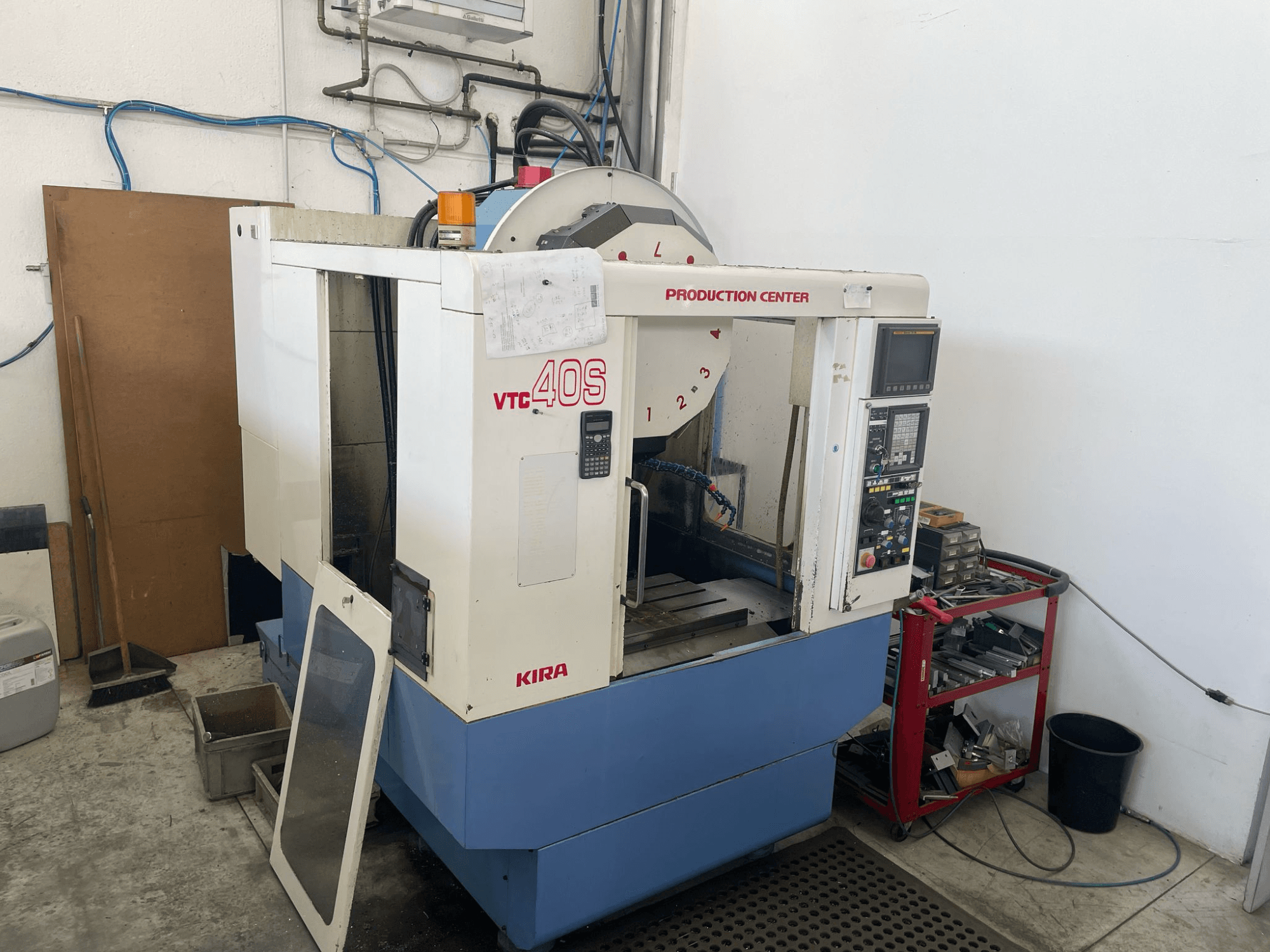 Centro de maquinação KIRA VTC 40S em vista lateral, com painel de controlo, porta aberta e ferramentas num carrinho de arrumação próximo.
