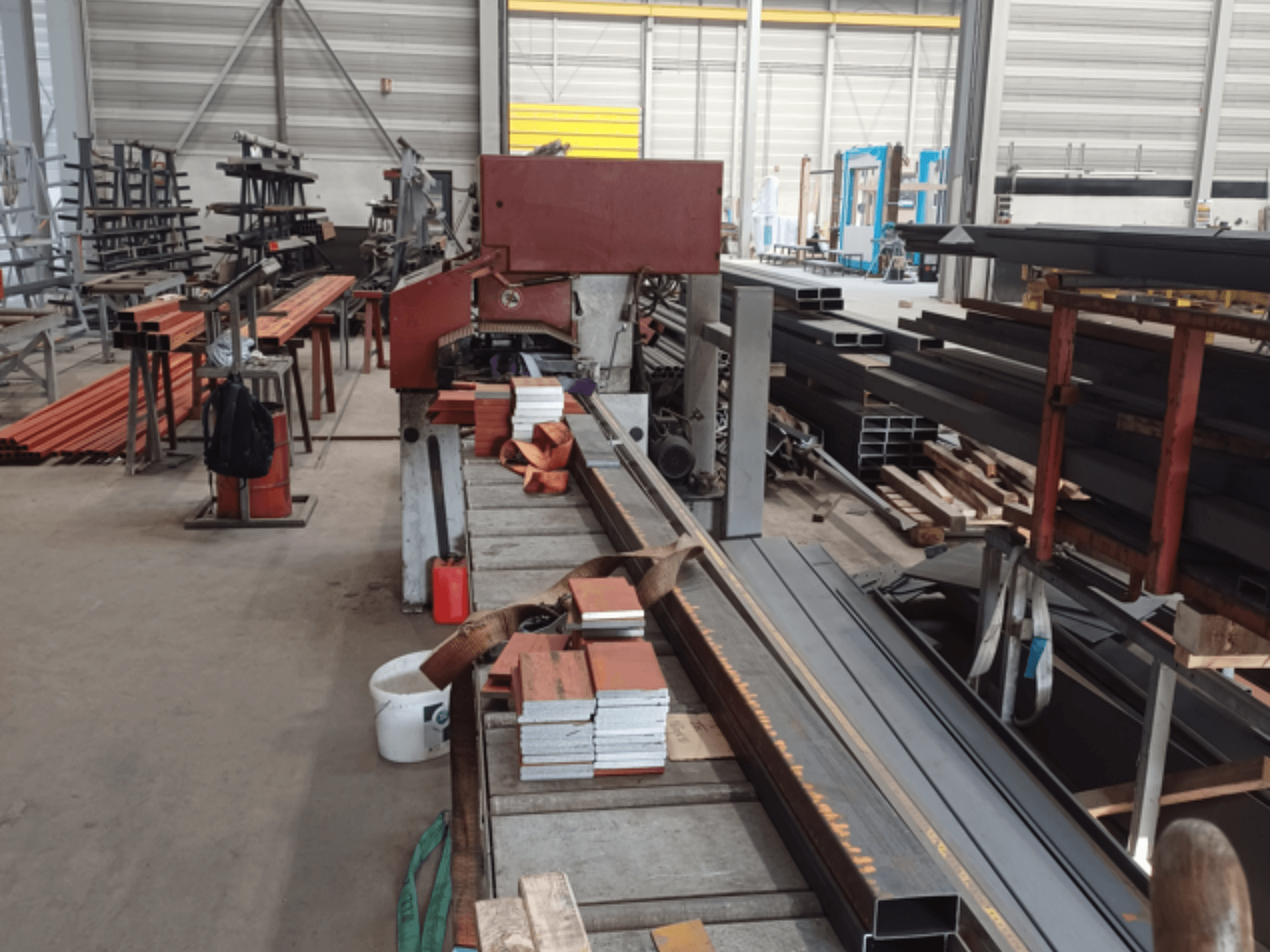 Oficina de fabrico de vários perfis metálicos, com uma máquina de serrar vermelha em primeiro plano e materiais empilhados.