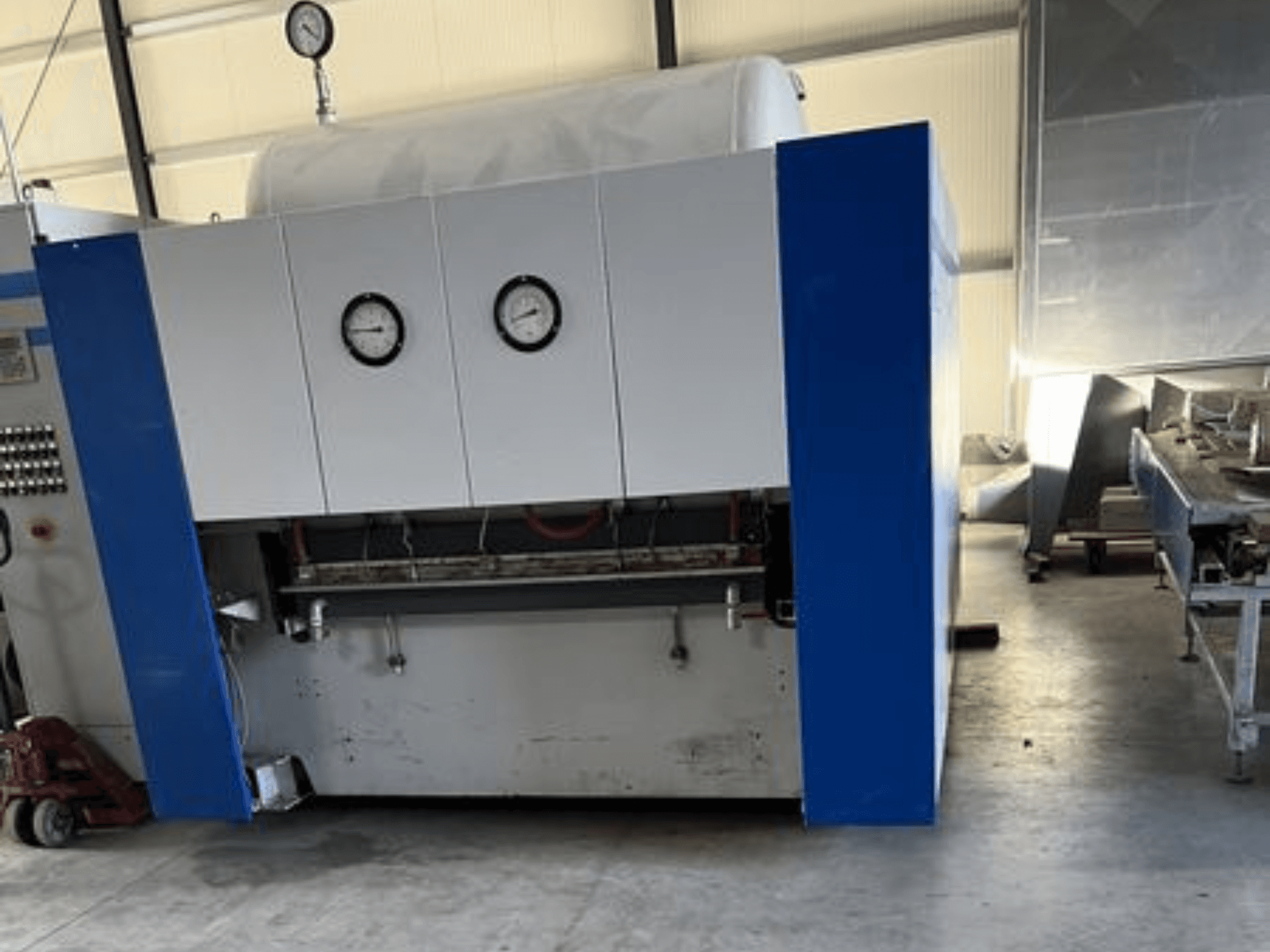 Máquina industrial azul e branca com dois manómetros, vista frontal, com um painel de controlo e equipamento adjacente.