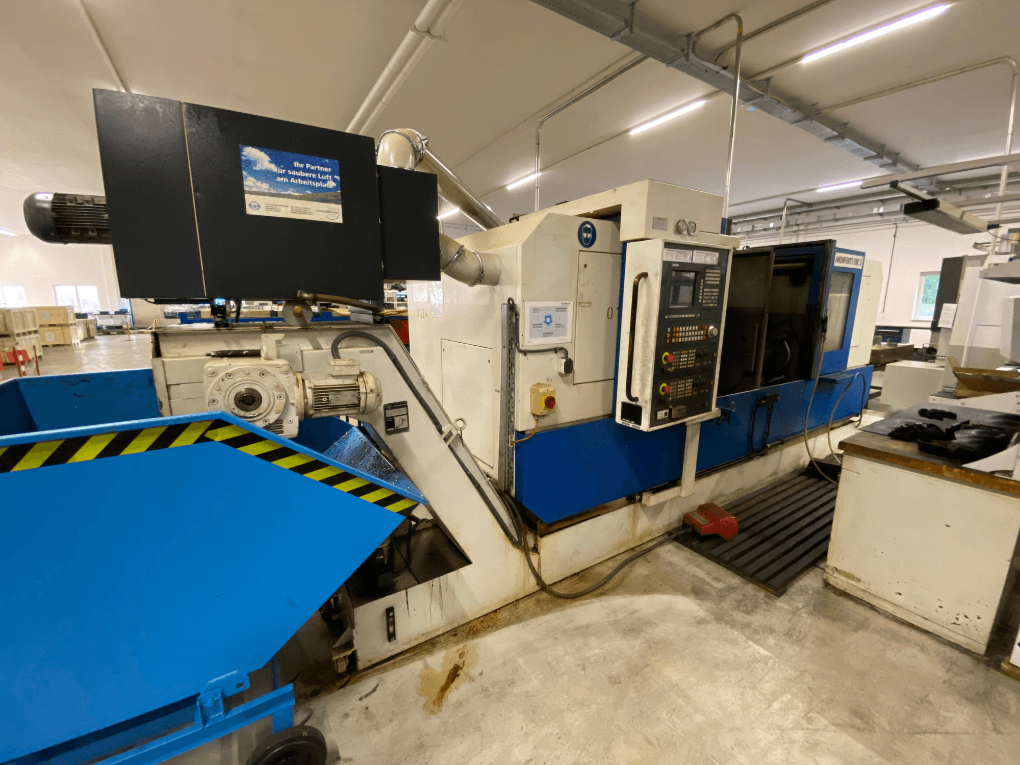 Centro de torneamento e fresagem Monforts DNC 5, vista lateral que mostra o painel de controlo, o tabuleiro azul para limalhas e detalhes da máquina num ambiente de oficina.