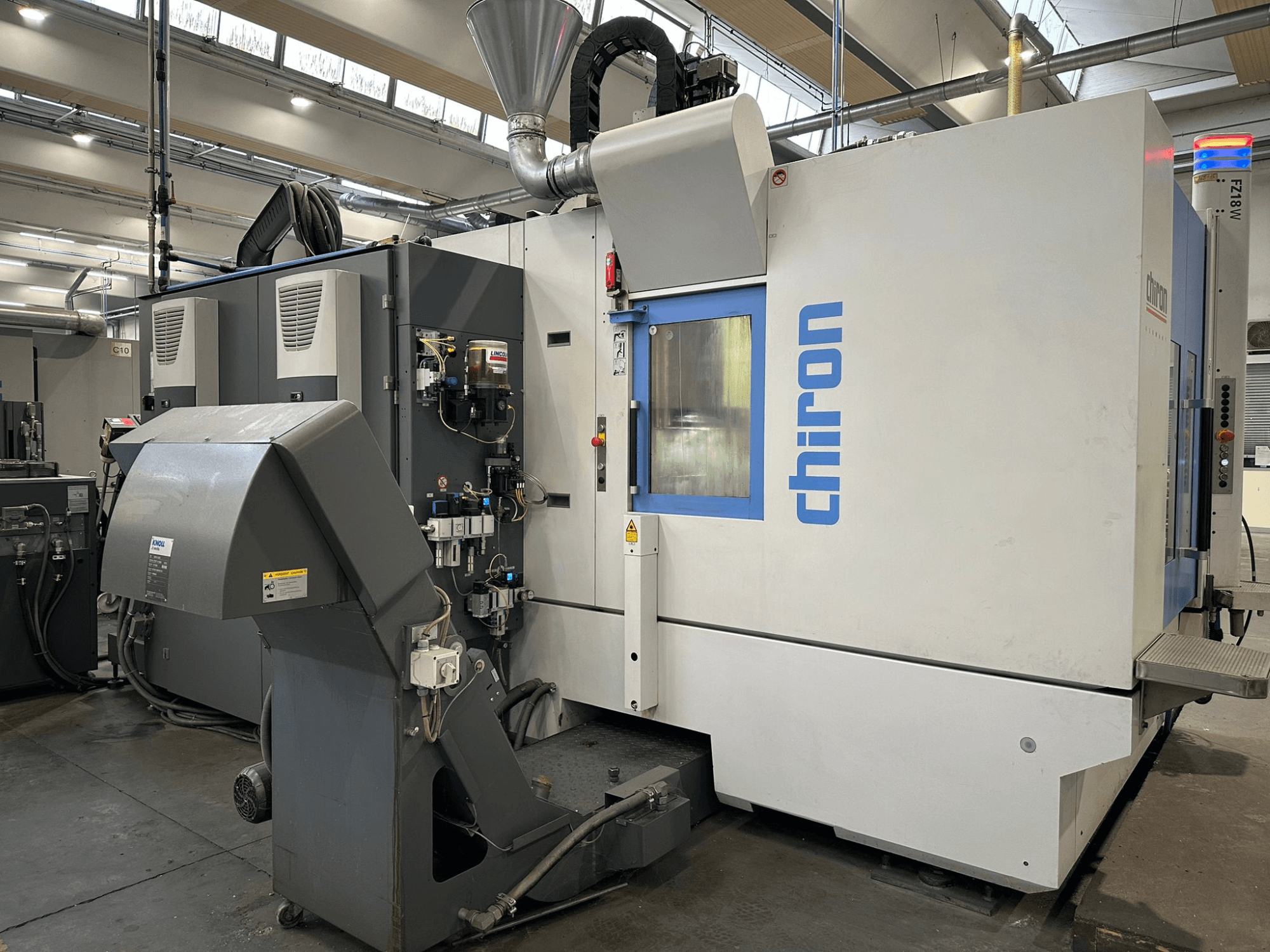 Máquina CNC Chiron em cinzento e branco, vista frontal, com comandos, tubos e um exaustor de poeiras.